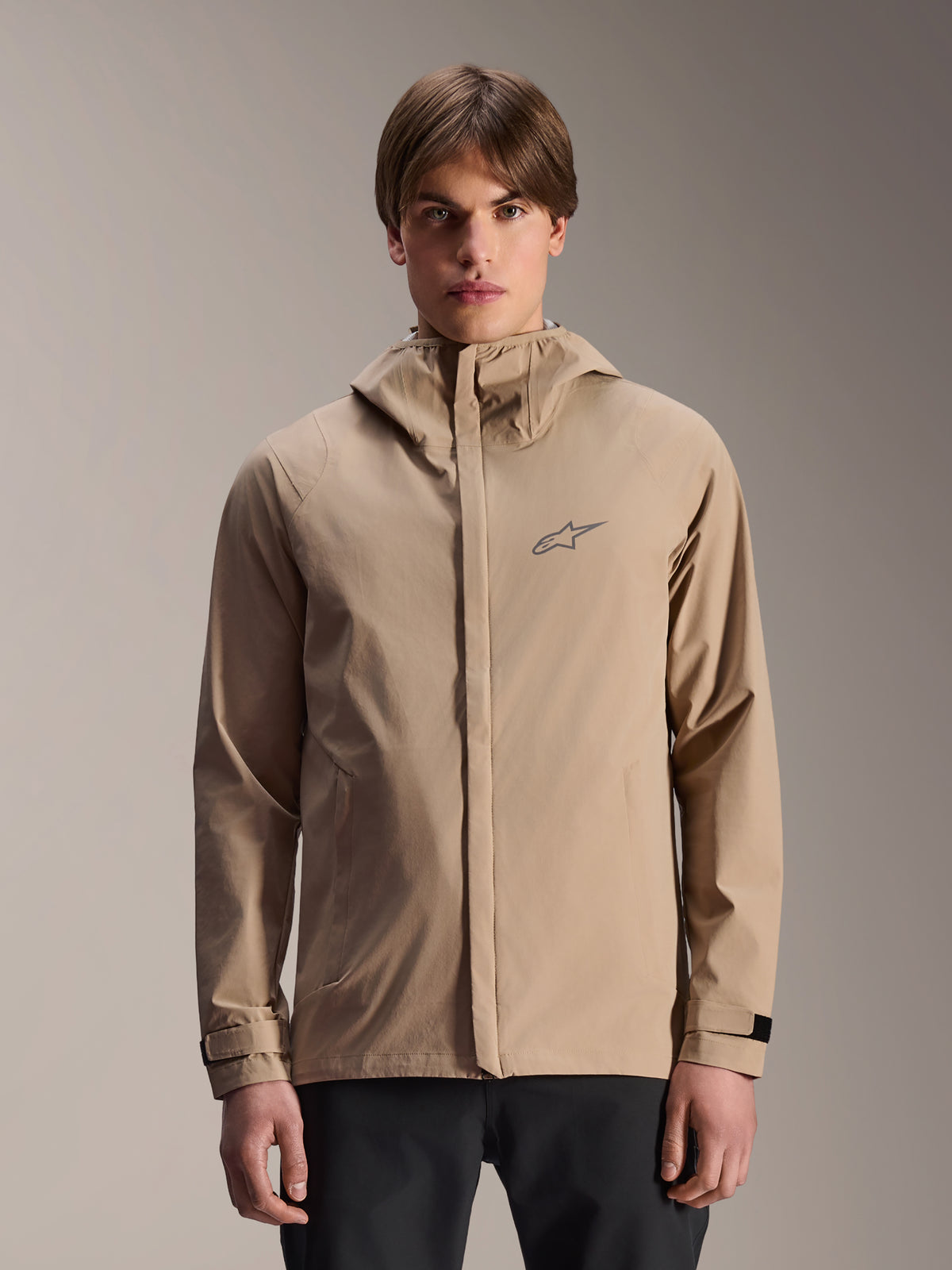 Alpinestars A-Dura Regenjas, lichtbruine mountainbike-regenjas met capuchon, gedragen door een model, met een klein logo op de borst, verstelbare manchetten en een verborgen ritssluiting, waterdichte fietskleding