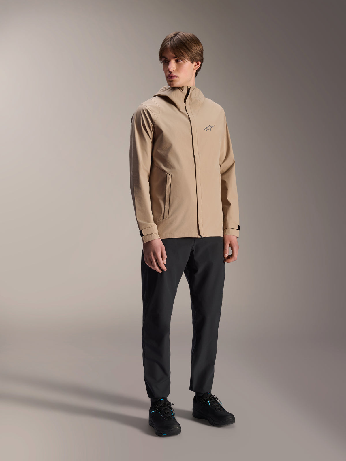 Mannelijk model draagt de Alpinestars A-Dura regenjas in lichtbruin, gecombineerd met een zwarte broek en schoenen, minimalistische outdoor mountainbike regenjas met capuchon en een klein logo op de borst.