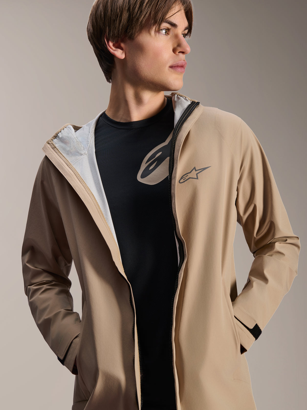 Mannelijk model draagt Alpinestars A-Dura regenjas, lichtbruin, geopend over een zwart T-shirt, witte mesh voering zichtbaar, veelzijdige weerbestendige fietsjas