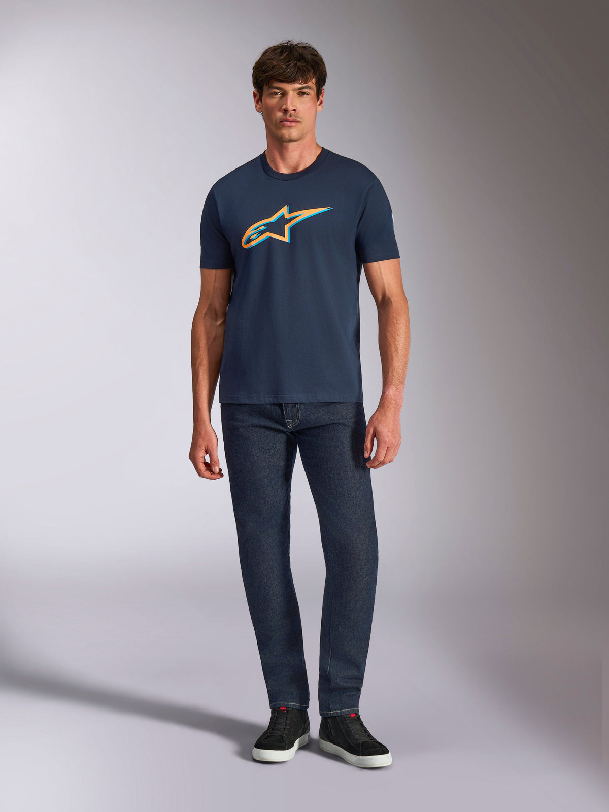 Alpinestars Ageless Shadow CSF Tee, T-shirt met korte mouwen, marineblauw, met een oranje en lichtblauw Ageless Shadow logo, gedragen door een model met een donkere denim jeans en zwarte sneakers, casual op motorsport geïnspireerde streetwear