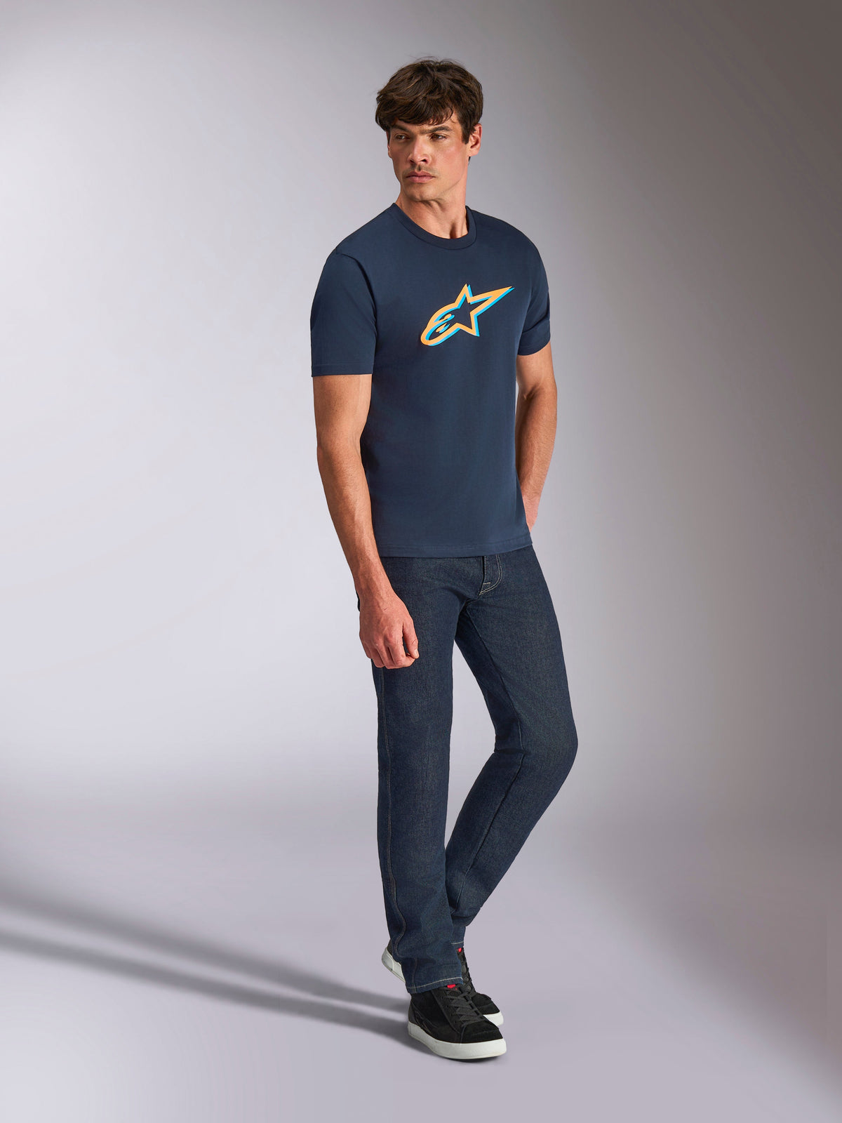 Alpinestars Ageless Shadow CSF Tee, T-shirt met korte mouwen, marineblauw met oranje en teal schaduwlogo, gedragen door een model met een donkere spijkerbroek en zwarte sneakers, casual motorsport streetwear