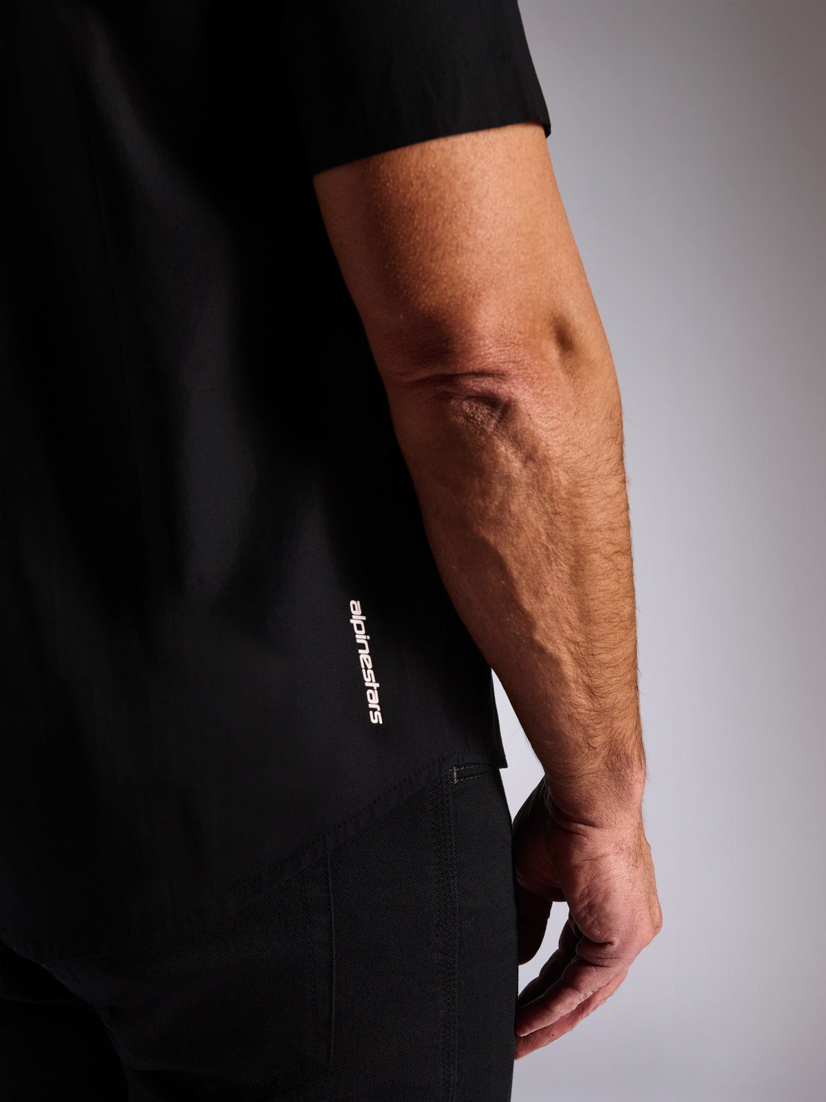 Alpinestars Strive Woven Shirt, Black short sleeve button-front shirt, vertical white Alpinestars logo on the side, close-up detail of the lower hem and brand logo, casual motorsport streetwear