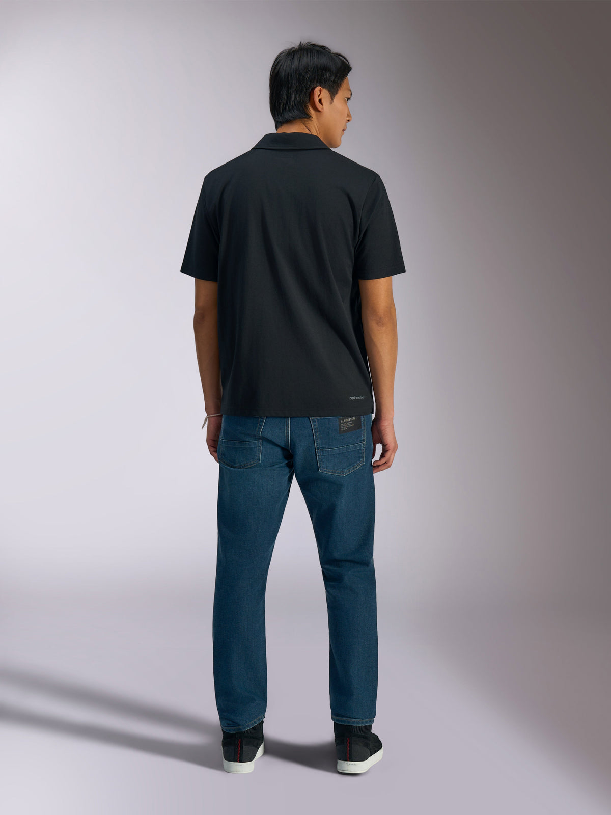 Alpinestars Impetus Sport Polo, zwart, achteraanzicht van poloshirt met korte mouwen met Alpinestars branding op de onderrug, gedragen met blauwe jeans, casual motorsport-geïnspireerde sportkleding