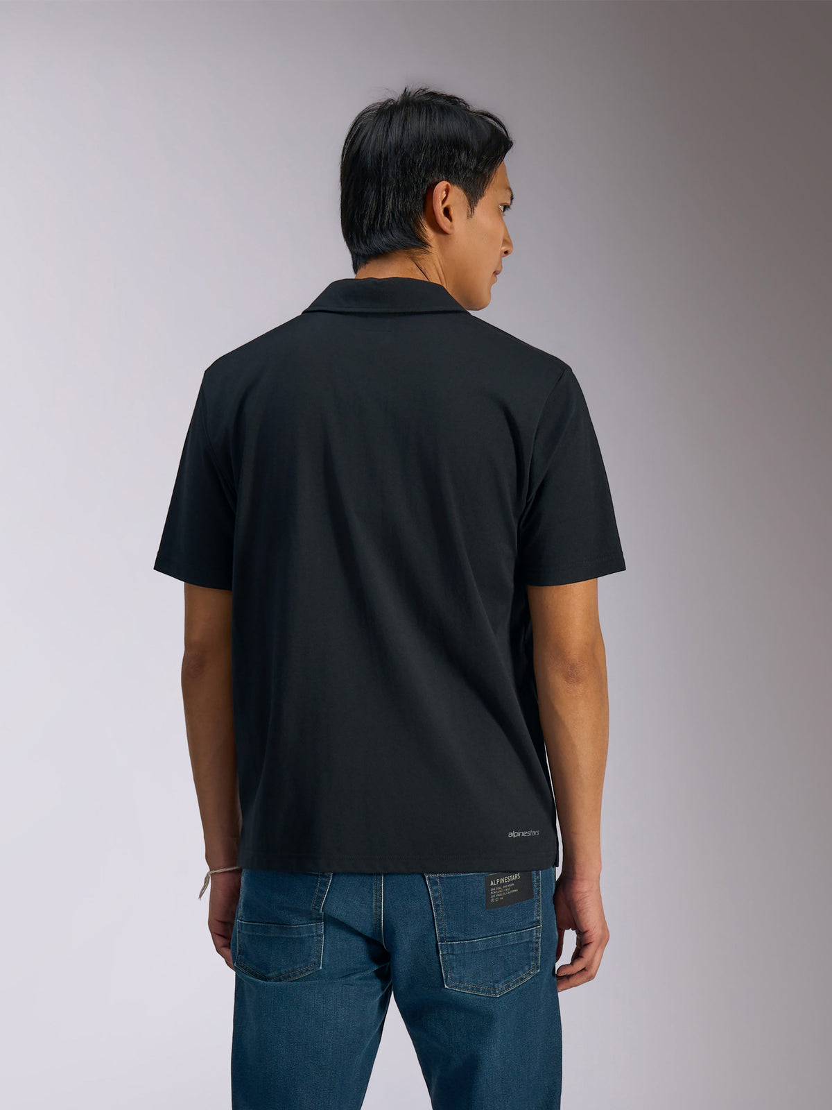 Alpinestars Impetus Sport Polo, zwart poloshirt met korte mouwen, achteraanzicht van een man die de polo draagt met een blauwe spijkerbroek, voorzien van een klein Alpinestars-woordmerk op de onderrug, casual op motorsport geïnspireerde sportkleding