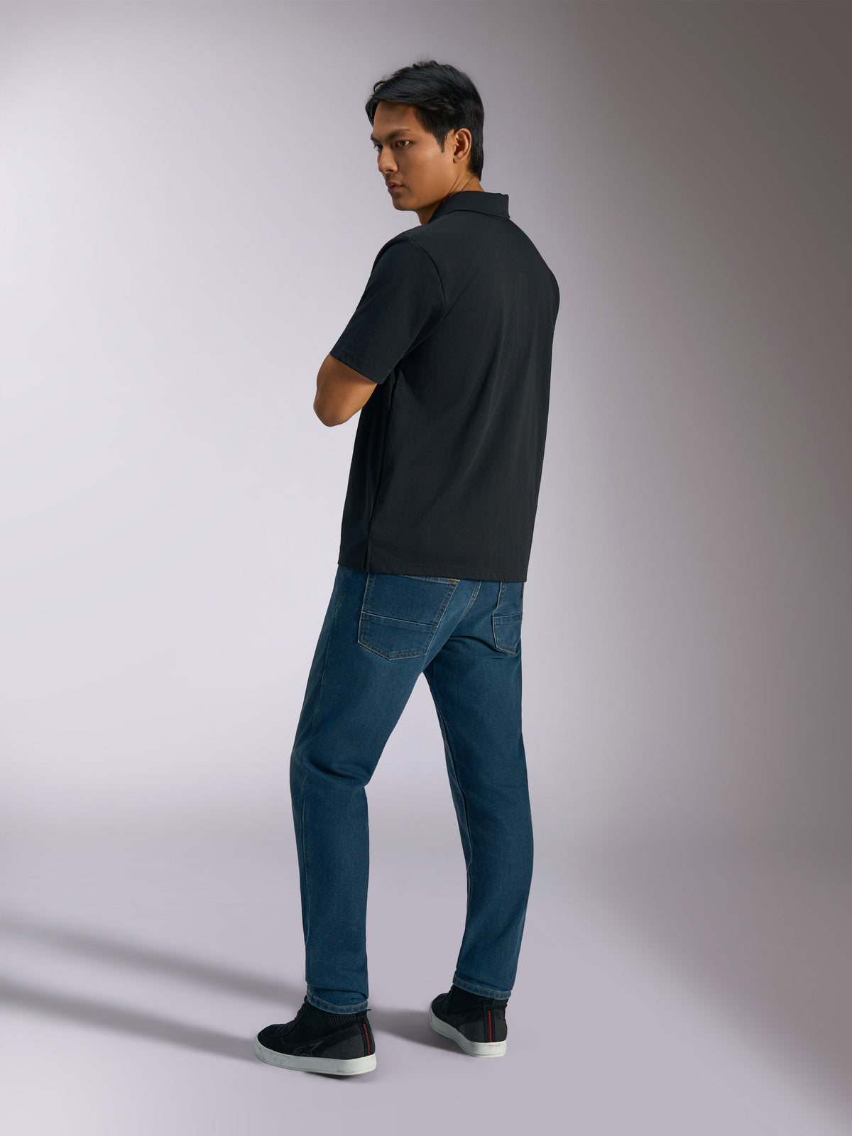 Alpinestars Impetus Sport Polo, zwart, poloshirt met korte mouwen met mock-mesh textuur en knoopsluiting met drie knopen, gedragen door een model met blauwe jeans en zwarte sneakers, casual streetwear geïnspireerd op de motorsport