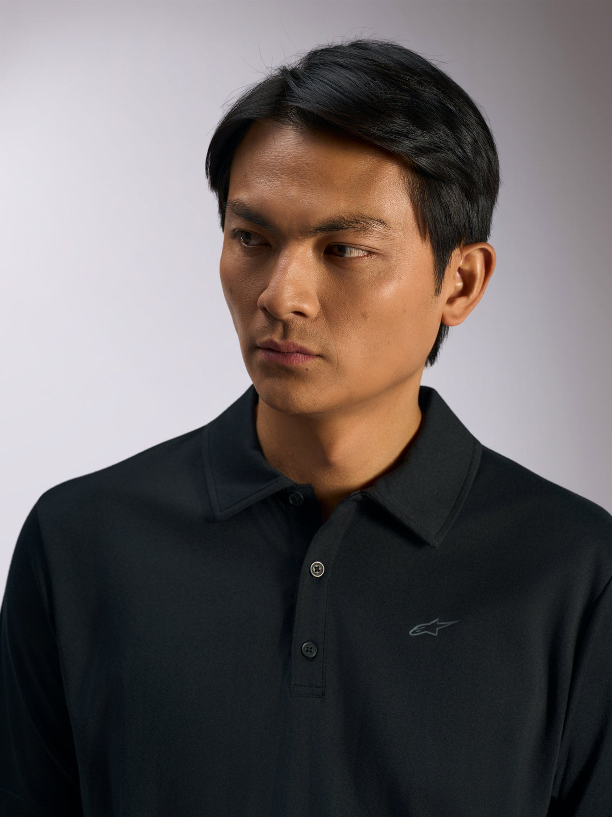 Alpinestars Impetus Sport Polo, zwart sportswear poloshirt, mock mesh-stof met een sluiting met drie knopen en een kraag van dezelfde stof, subtiel Astars-logo op de borst, casual motorsport lifestyle-kleding, close-up van een persoon die de polo draagt