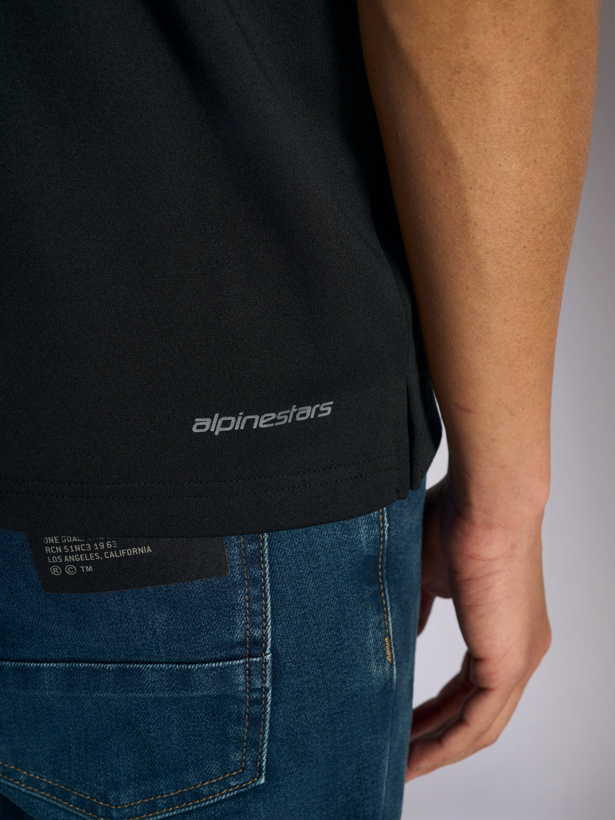 Alpinestars Impetus Sport Polo, detailweergave van de zoom aan de onderkant en zijsplit, zwart met grijze Alpinestars woordmerk-branding, casual motorsport-geïnspireerde sportkleding