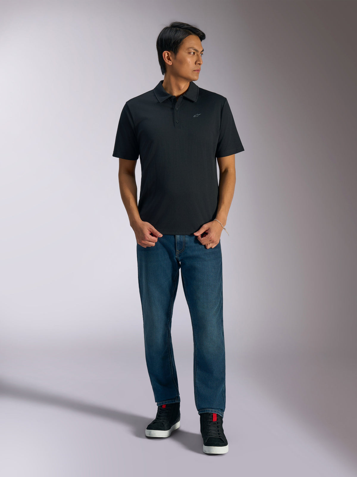 Alpinestars Impetus Sport Polo, zwart poloshirt met korte mouwen, voorzien van een knoopsluiting met drie knopen en een grijs Astars-logo op de borst, gedragen door een man met een blauwe jeans en zwarte hoge sneakers, casual motorsport-geïnspireerde sportkleding