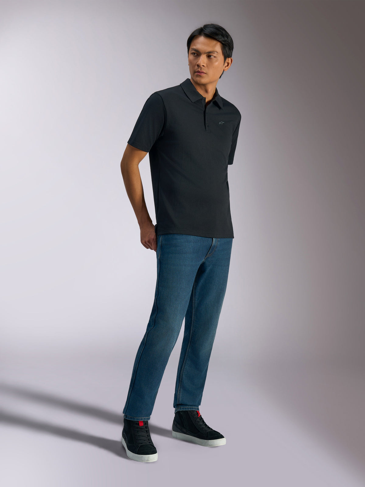 Alpinestars Impetus Sport Polo, poloshirt met korte mouwen, zwart, voorzien van een subtiel logo op de borst en een sluiting met drie knopen, gedragen door een model met blauwe jeans en zwarte sneakers, casual motorsport-geïnspireerde lifestylekleding