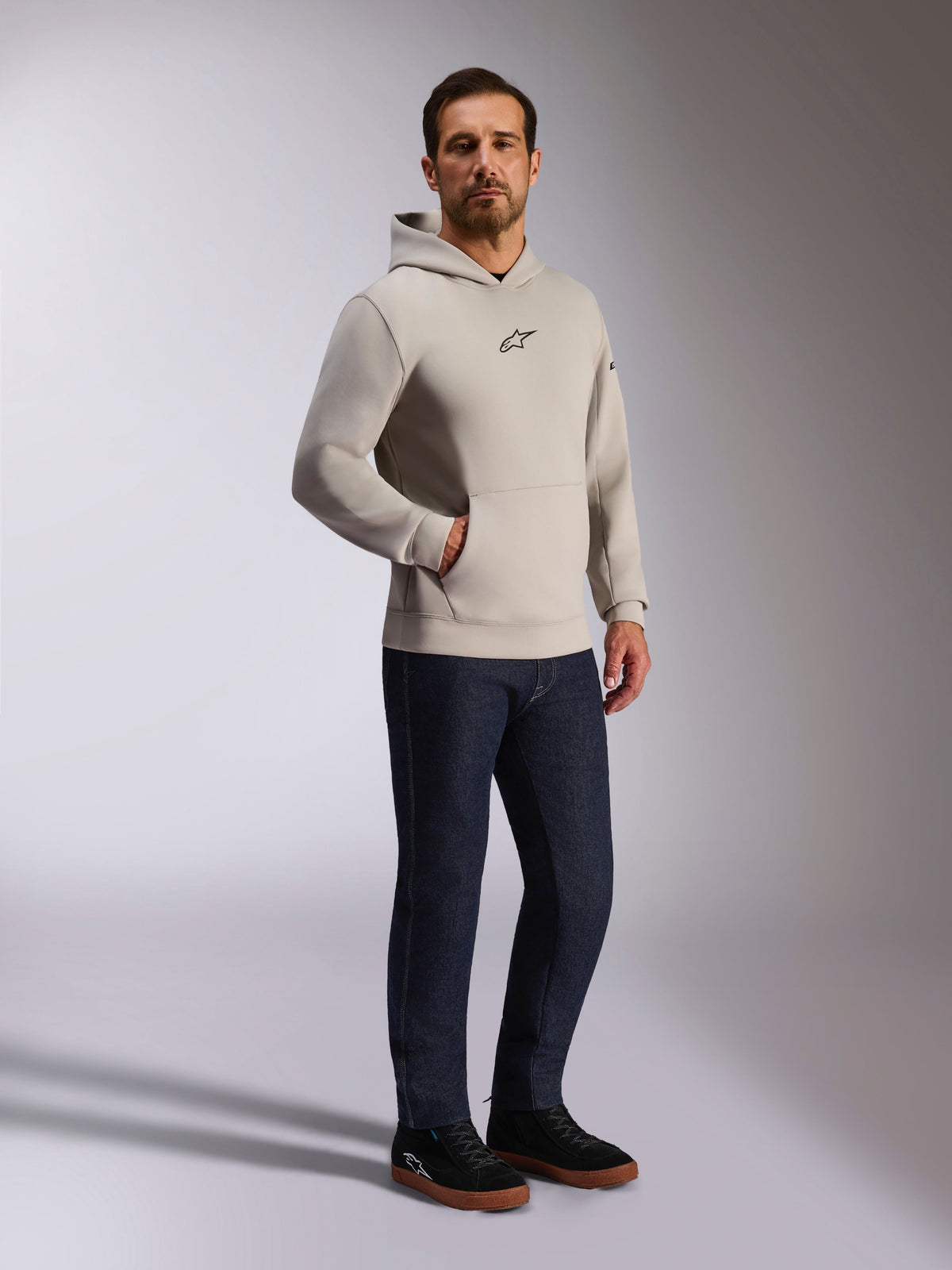 Alpinestars Insight Hoodie, casual streetwear, Cement gray with black Astars logo on chest, pullover design with kangaroo pocket, worn with dark blue jeans and black sneakers, modern motorsport-inspired style