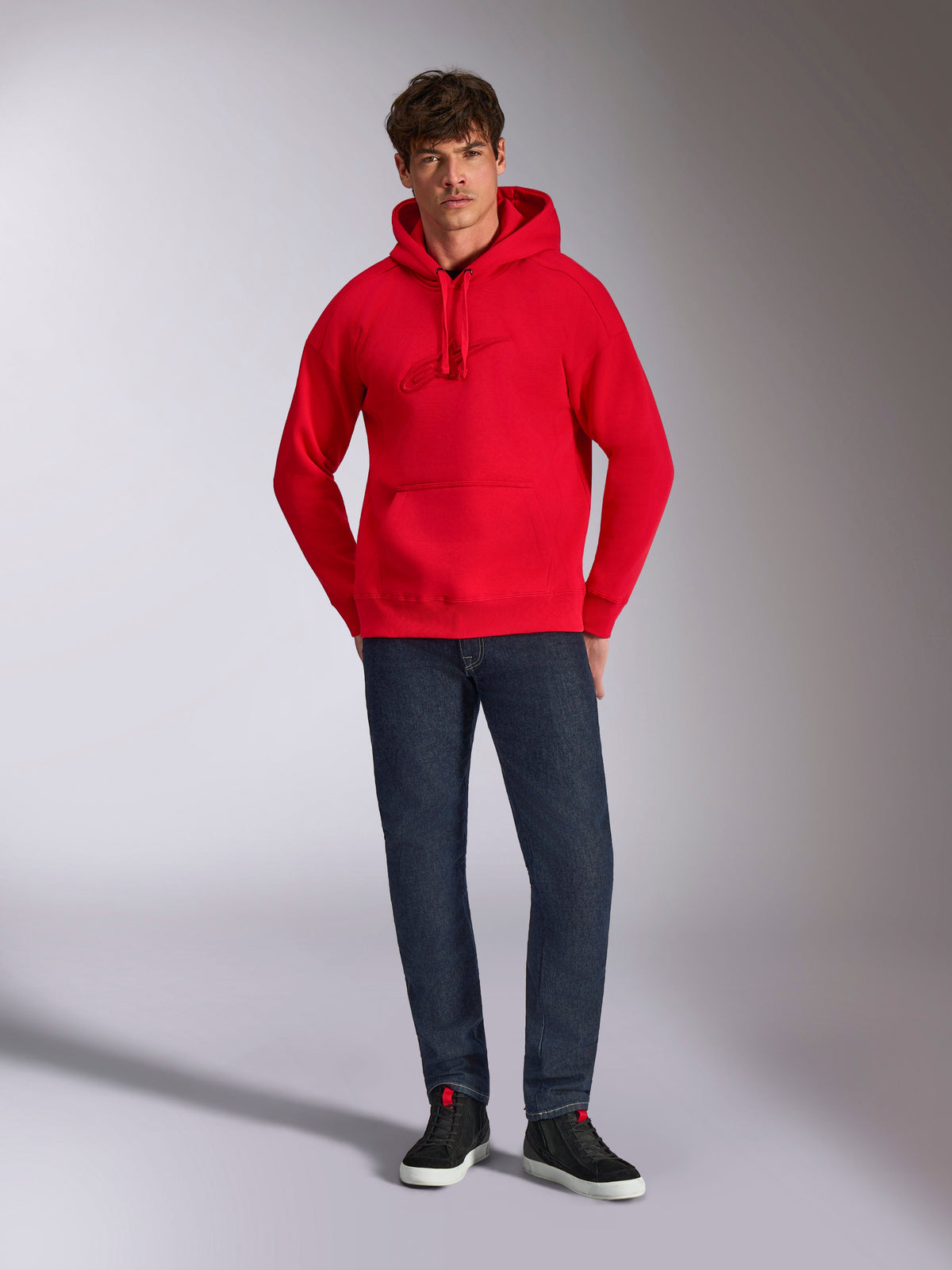 Alpinestars Emergent Oversized Hoodie, rood, met ton-sur-ton 3D-geborduurd Astars-logo op de borst, kangoeroezak en capuchon met trekkoord, gedragen door een model met donkerblauwe jeans en zwarte sneakers, casual lifestyle streetwear