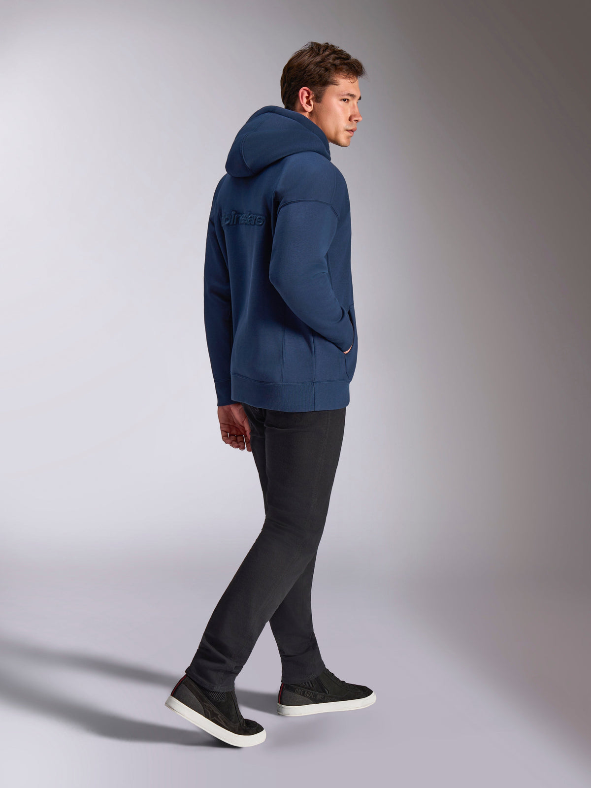 Alpinestars Emergent Oversized Hoodie in marineblauw, achteraanzicht met 3D-geborduurd Alpinestars-woordlogo, oversized fleece pasvorm met capuchon met trekkoord, gedragen met een zwarte broek, casual motorsport-geïnspireerde streetwear