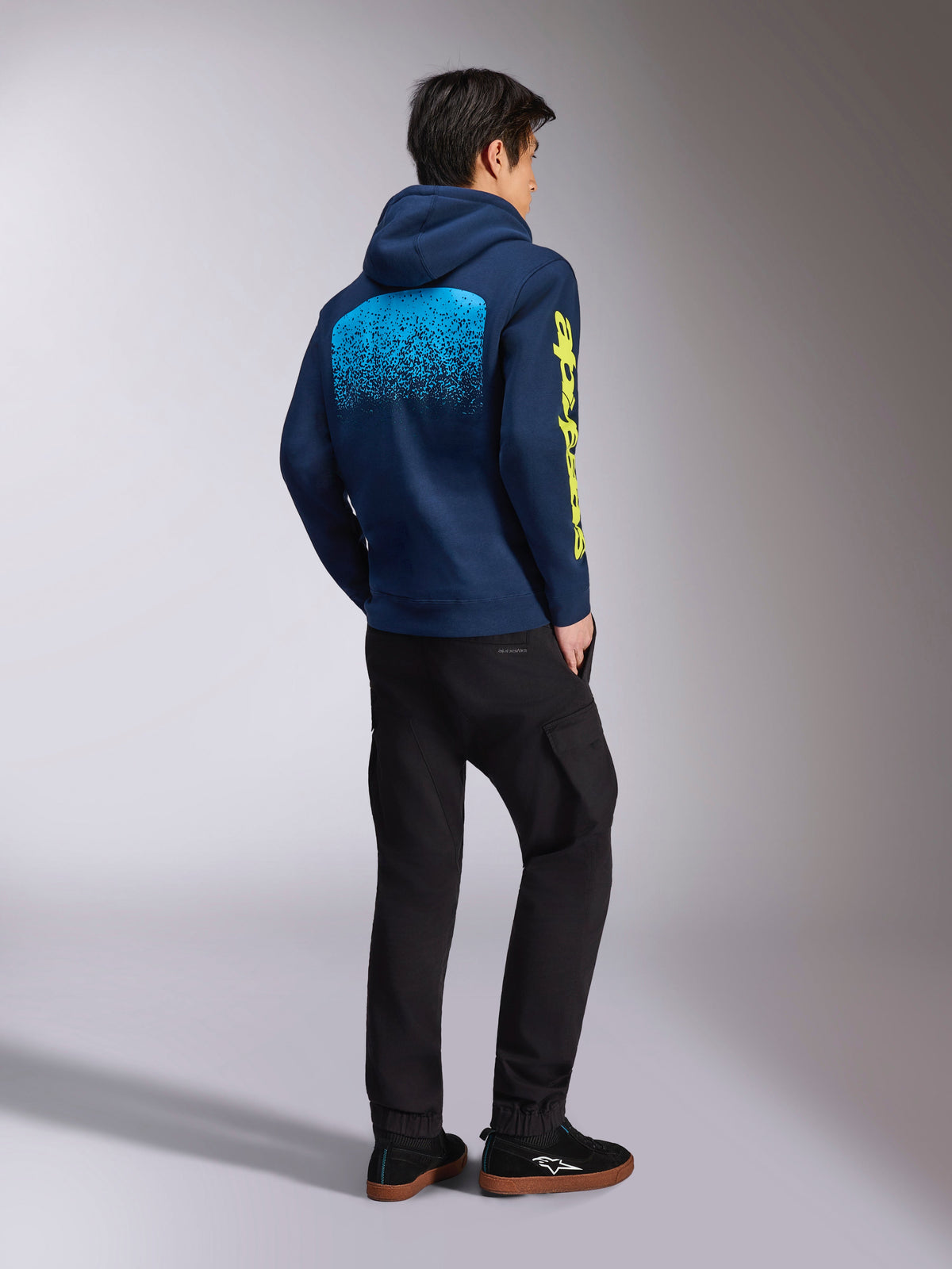Alpinestars Obsessed Hoodie, navy blauwe casual sweatshirt, achteraanzicht met lichtblauwe MX-verloopgraphic en geel Alpinestars-logo op de mouw, gedragen met een zwarte broek en sneakers