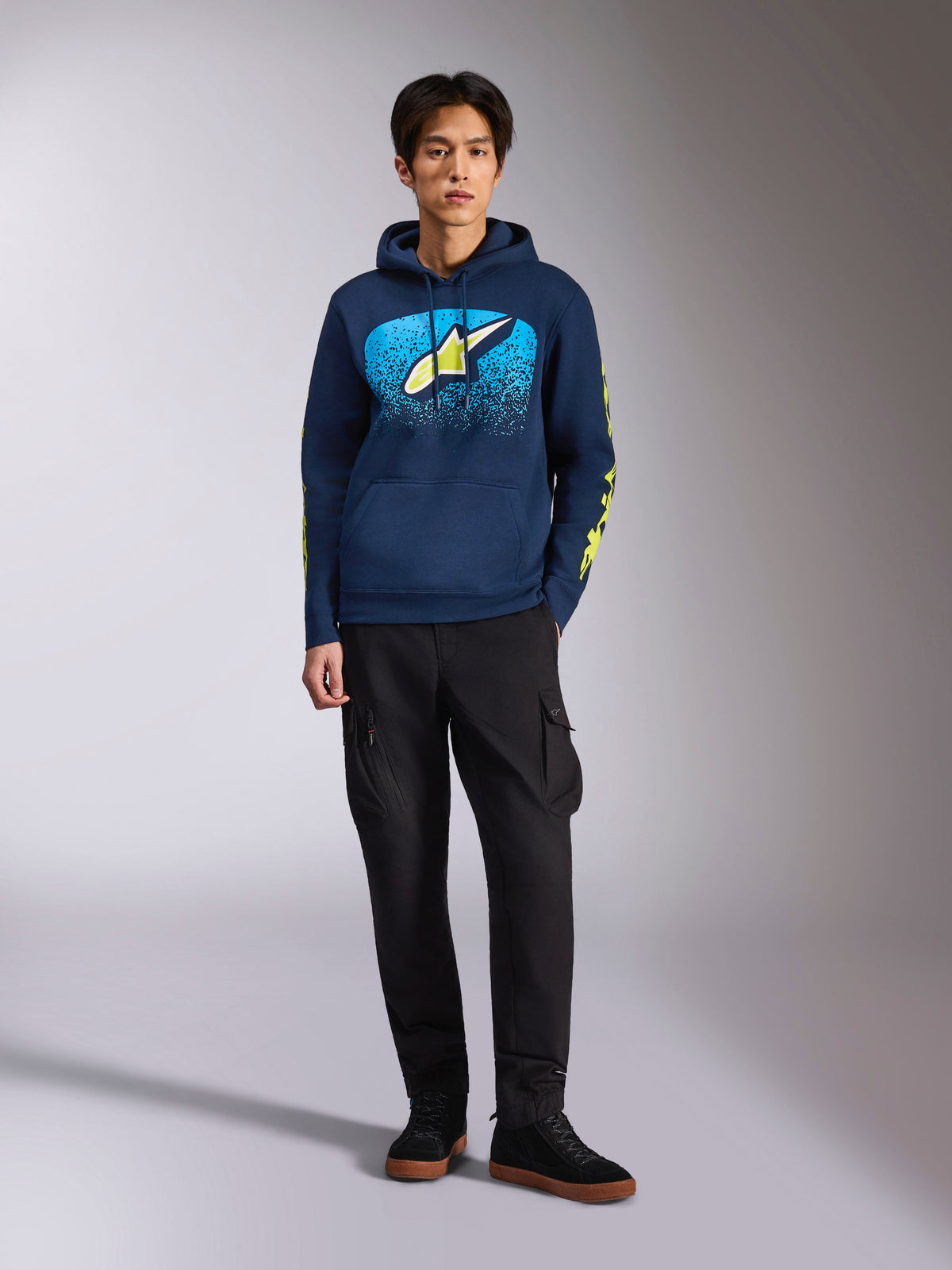 Alpinestars Obsessed Hoodie, navyblauwe pullover hoodie, Astars-logo op de borst met blauw MX-verloop en limoengele ster, limoengele Alpinestars-woordlogo's op de mouwen, gedragen door een model met zwarte cargobroek en zwarte sneakers, casual motorsport-geïnspireerde streetwear