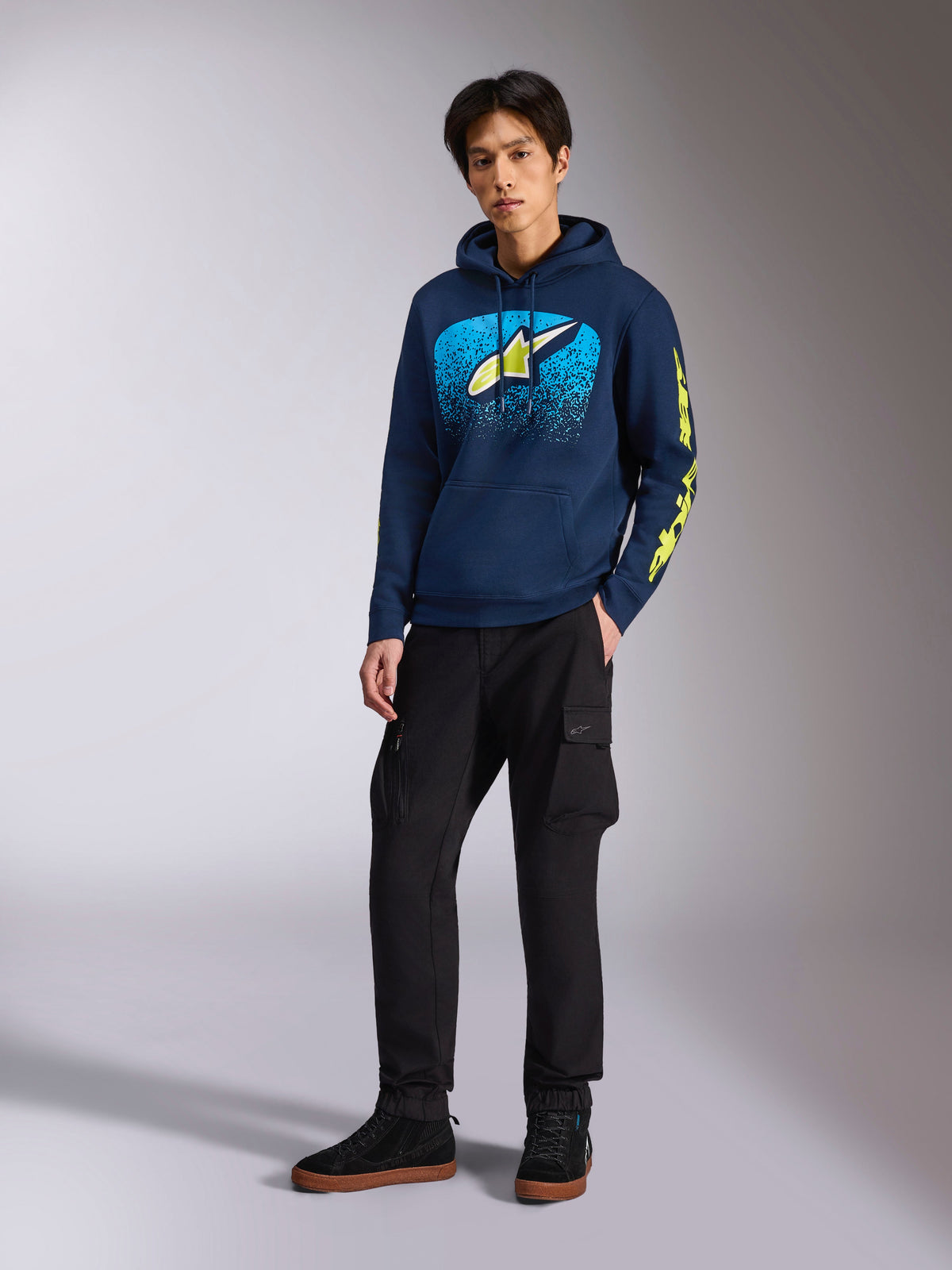 Alpinestars Obsessed Hoodie in Navy, casual pullover fleece met lichtblauwe en limoengele graphic op de borst, limoengele Alpinestars-branding op de mouw, gedragen door een model met een zwarte cargobroek en zwarte sneakers, op motorsport geïnspireerde streetwear