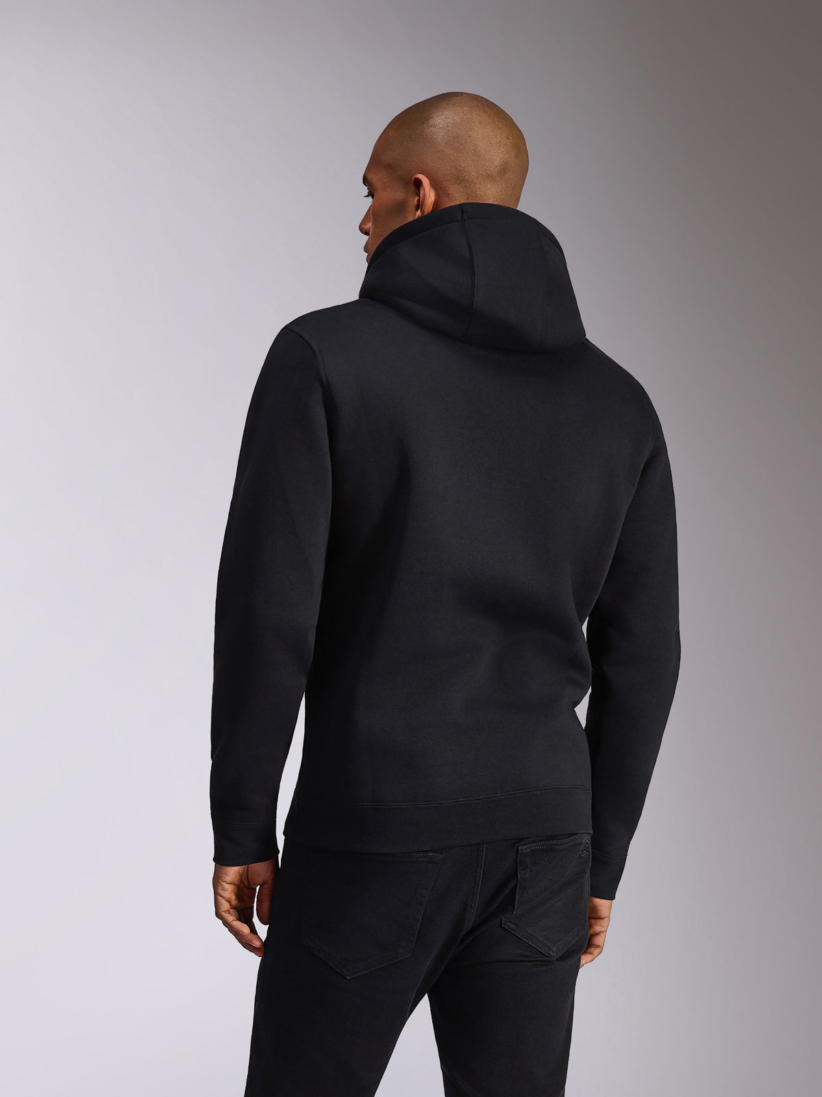 Alpinestars Ellipsoid Hoodie, zwart, achteraanzicht van een persoon die een fleece hoodie draagt, lange mouwen met geribde manchetten en zoom, gecombineerd met een zwarte jeans, casual op de motorsport geïnspireerde sportkleding