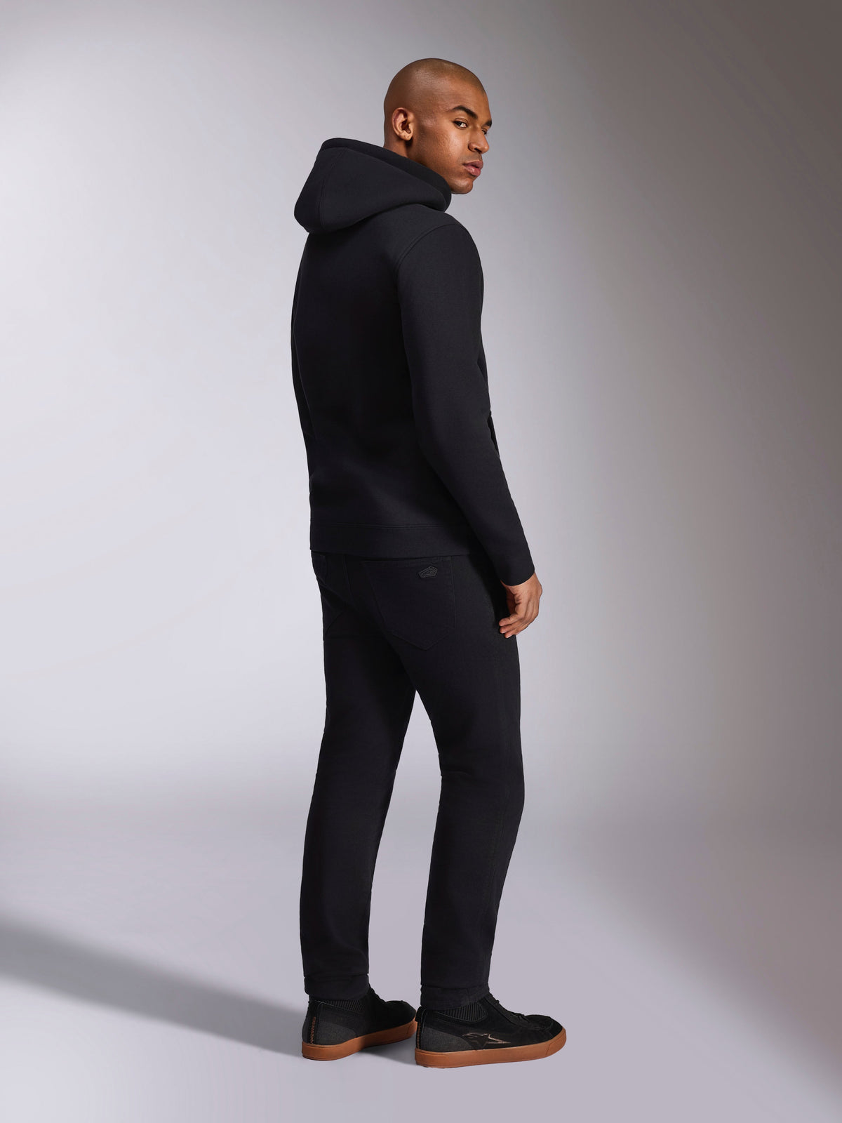 Alpinestars Ellipsoid Hoodie, zwarte casual streetwear, achteraanzicht op mannelijk model, fleece pullover met capuchon, geribde manchetten en zoom, gedragen met zwarte broek en sneakers, modern op de motorsport geïnspireerd ontwerp