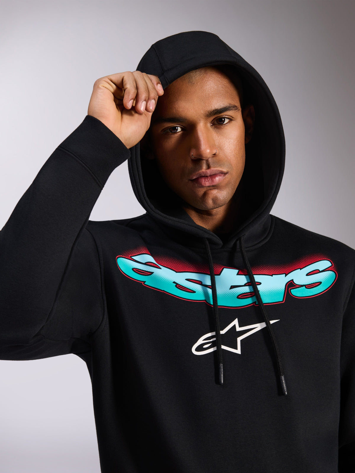 Alpinestars Ellipsoid Hoodie, zwarte pullover fleece hoodie, met gestileerd Astars-woordmerk in blauwgroen en rood kleurverloop en wit logo op de borst, gedragen door model met de capuchon op, casual motorsport-geïnspireerde streetwear
