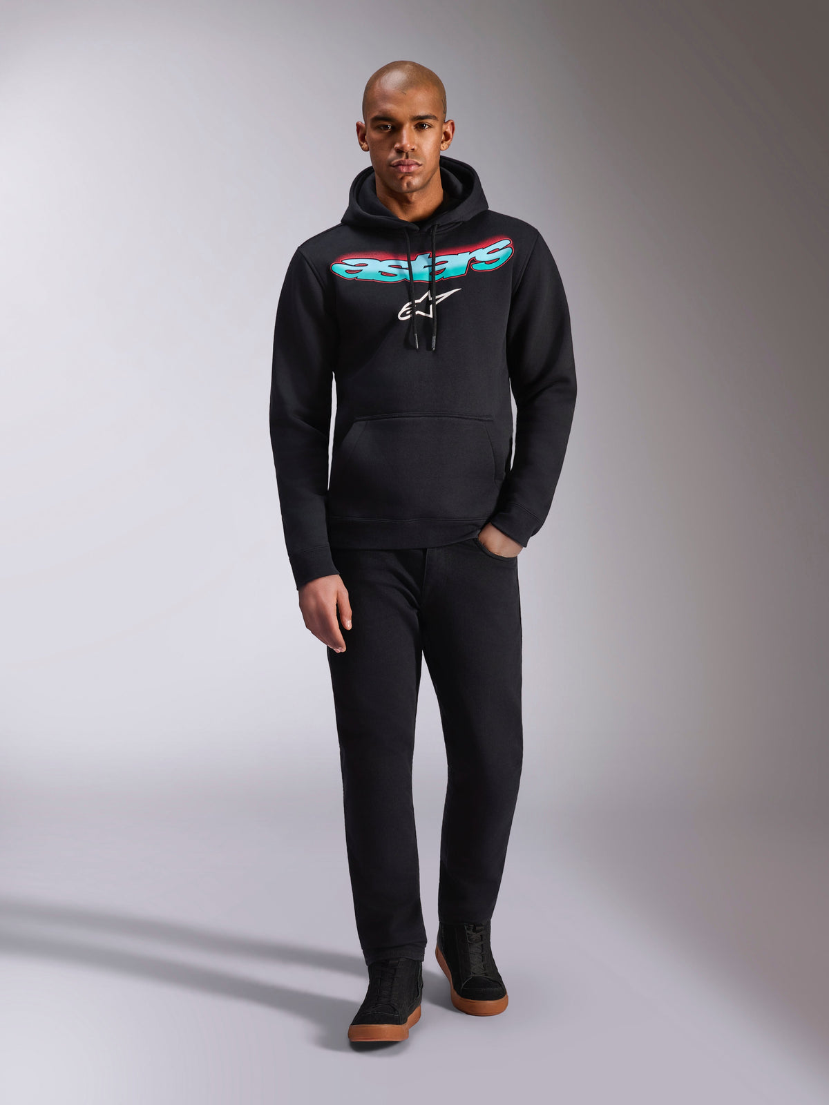 Alpinestars Ellipsoid Hoodie, zwart, pullover fleece met een gebogen Astars-tekst met een rood en blauwgroen kleurverloop en wit sterlogo, gedragen door een model met zwarte broek en sneakers, casual motorsport streetwear