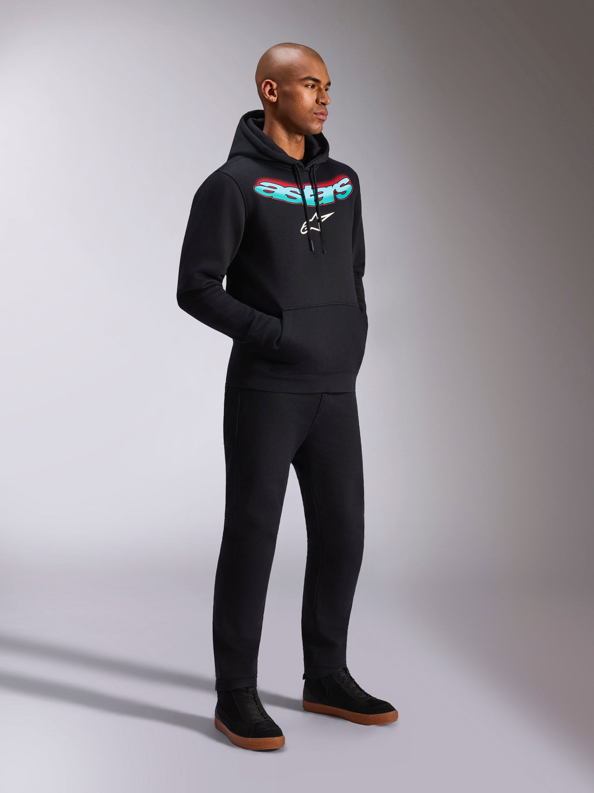 Alpinestars Ellipsoid Hoodie, zwarte fleece hoodie met capuchon, voorzien van een gebogen Astars-woordmerk met een groenblauw en rood kleurverloop en een wit sterlogo, gedragen door een model met een zwarte broek, casual motorsport-geïnspireerde streetwear