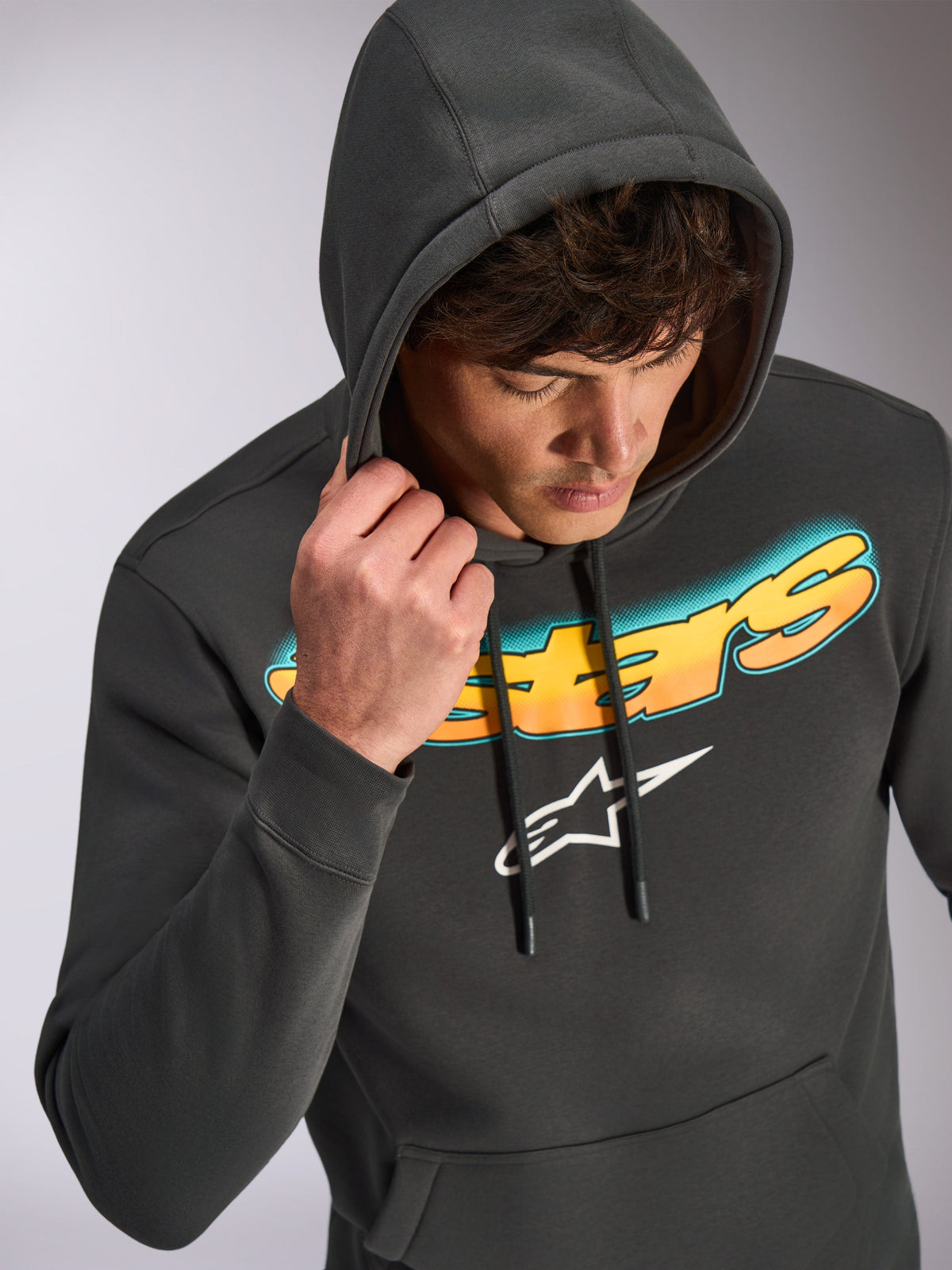 Alpinestars Ellipsoid Hoodie, gunmetal grijs, casual pullover hoodie met een Astars-woordmerk in een kleurverloop van cyaan naar oranje en een wit logo, gedragen door een model met de capuchon op, op motorsport geïnspireerde streetwear