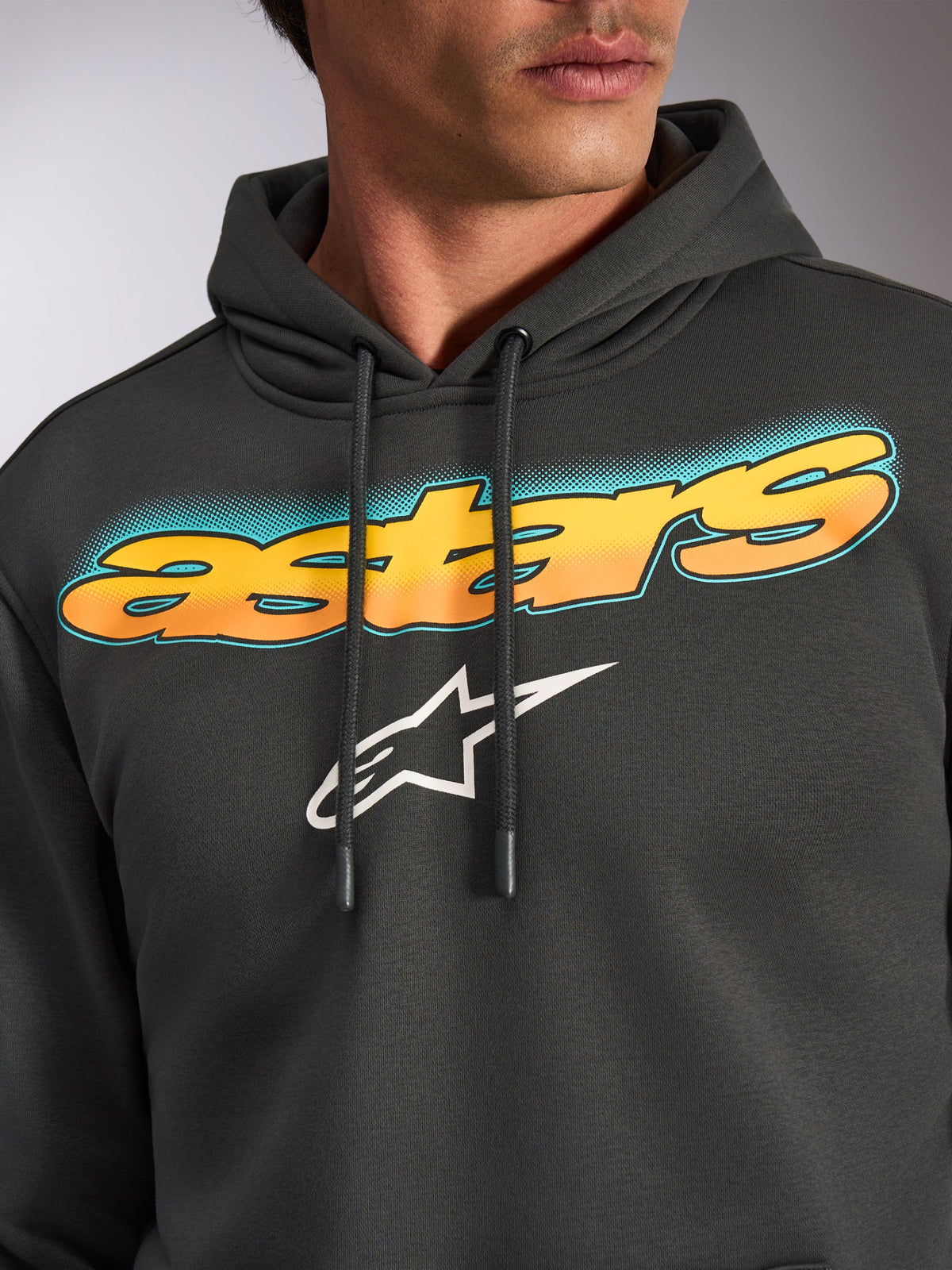 Alpinestars Ellipsoid Hoodie, Gunmetal grijs, close-up van de borst met gebogen 'astars' tekst met geel-oranje kleurverloop en teal accenten, wit Alpinestars ster-logo, casual motorsport-geïnspireerde streetwear fleece