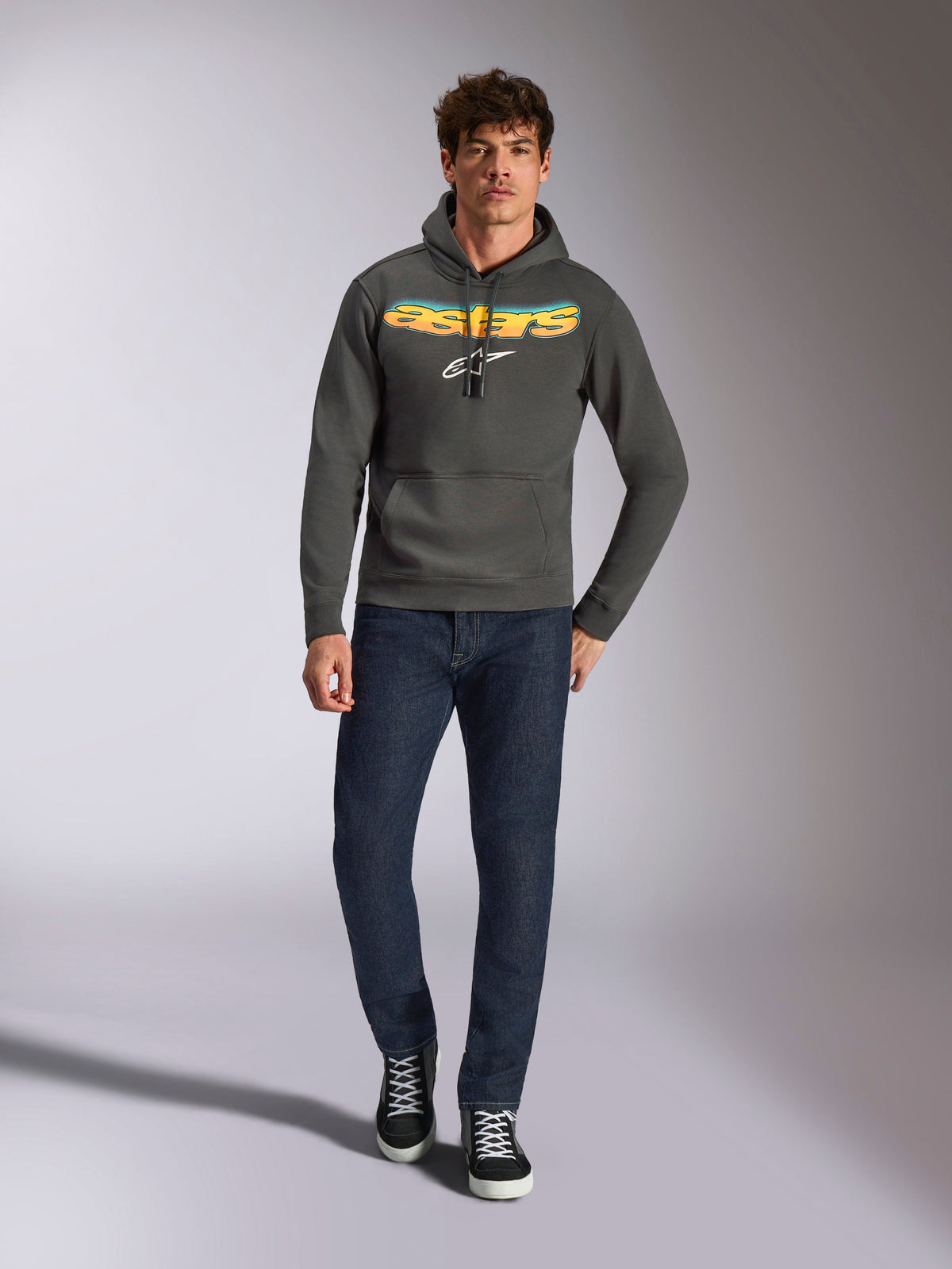 Alpinestars Ellipsoid Hoodie, casual sportkleding, gunmetal grijs met oranje en geel kleurverloop Astars-logo en wit symbool, capuchon met trekkoord, gedragen door een mannelijk model met donkerblauwe jeans en zwarte sneakers, moderne op motorsport geïnspireerde streetwear