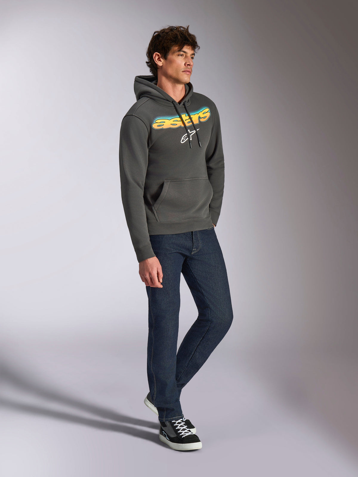 Alpinestars Ellipsoid Hoodie, gunmetal grijze pullover fleece, gebogen Astars-woordmerk aan de voorzijde met een blauw en geel kleurverloop en wit logo, gestyled met een donkere jeans en zwarte sneakers, casual streetwear.
