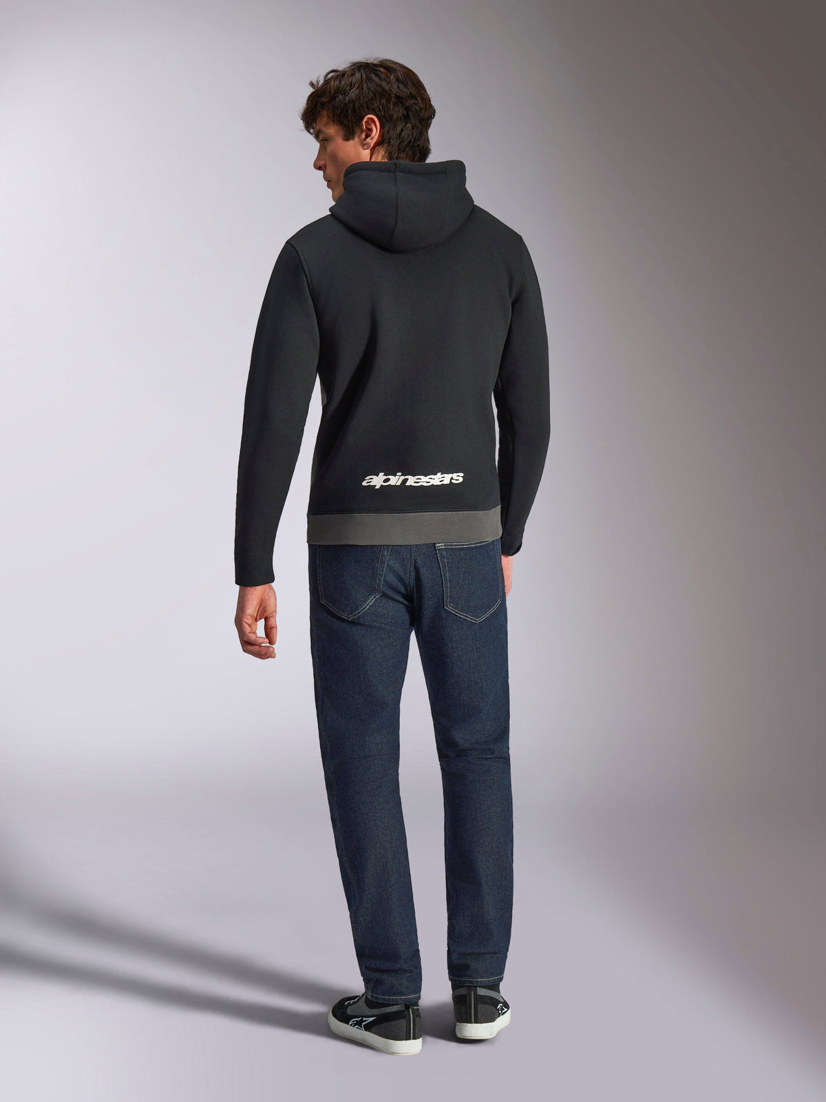 Alpinestars Parallel Hoodie, achteraanzicht van een man in een zwart en gunmetal kleurgeblokte fleece trui, wit Alpinestars woordlogo op de onderrug, gecombineerd met een donkerblauwe jeans, casual op motorsport geïnspireerde streetwear