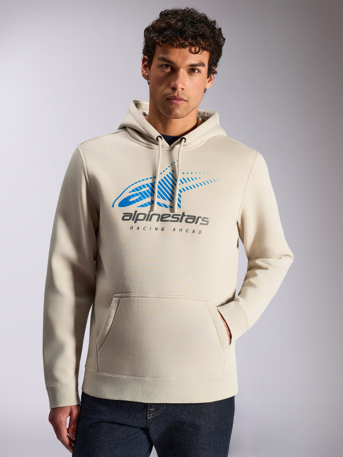 Alpinestars Pulse Hoodie, casual streetwear, zandkleur, blauw en grijs Astars Combo Logo op de voorzijde met speed line fade en 'Racing Ahead' tekst, capuchon met trekkoord, kangoeroezak, gedragen door een model met een donkere jeans