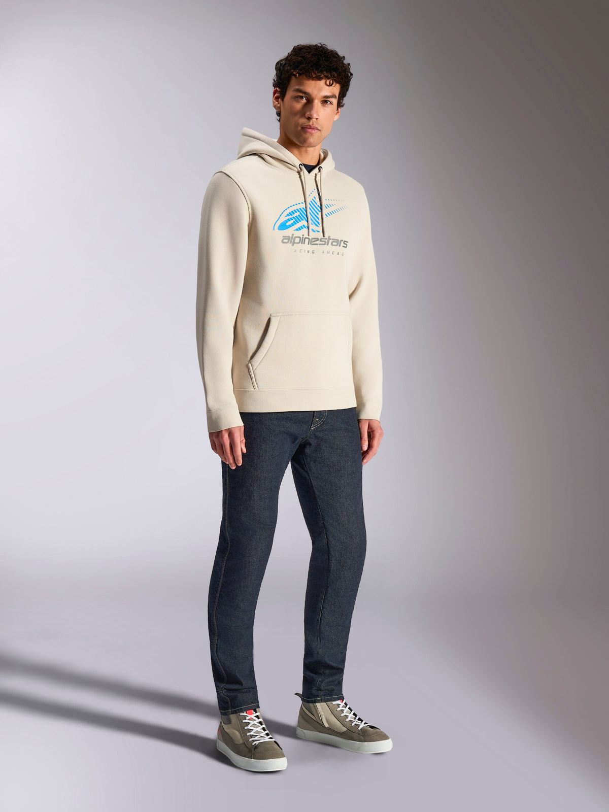Alpinestars Pulse Hoodie, Off White casual streetwear, fleece pullover met capuchon voorzien van een blauw en grijs Astars-logo met speed line fade, kangoeroezak, gedragen door model met donkere denim jeans en high-top sneakers.