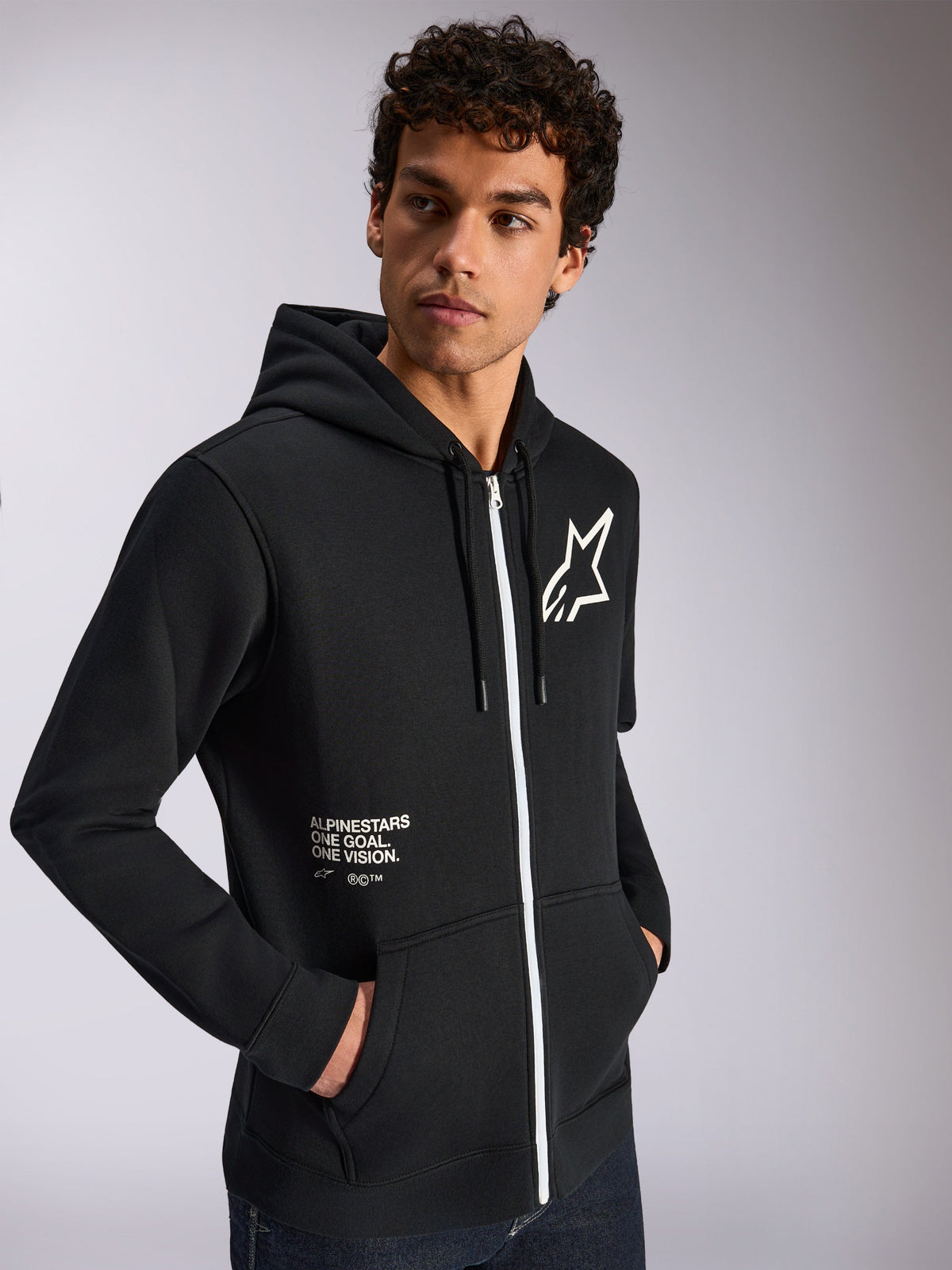 Alpinestars Transverse Hoodie, casual sportkleding, zwart met wit ritsdetail, capuchon met trekkoord, Alpinestars ster-logo op de borst, 'One Goal One Vision' graphic, gedragen door een mannelijk model met donker krullend haar