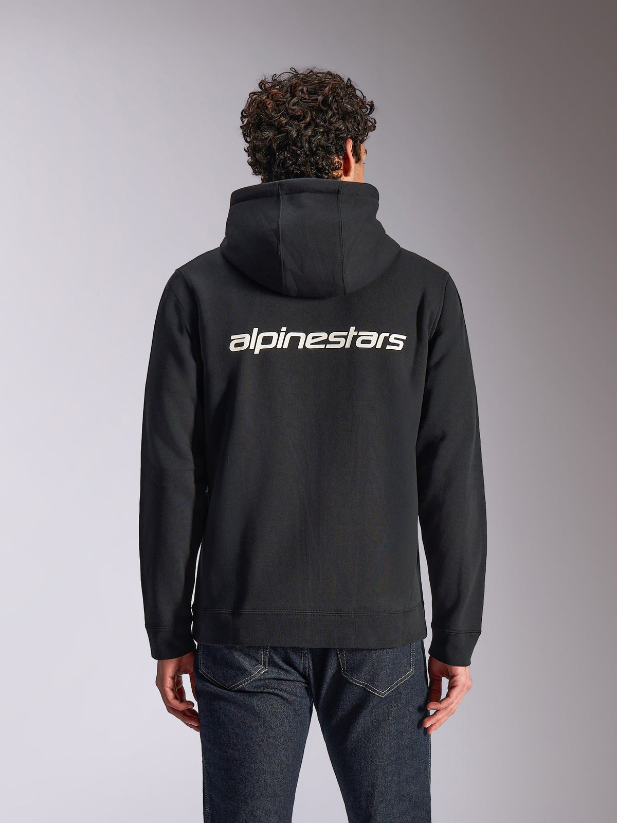 Alpinestars Transverse Hoodie, casual streetwear, zwart met wit Alpinestars-woordlogo op de rug, fleece hoodie met rits aan de voorkant, lange mouwen, gedragen door een model met donkere spijkerbroek, modern op motorsport geïnspireerd design