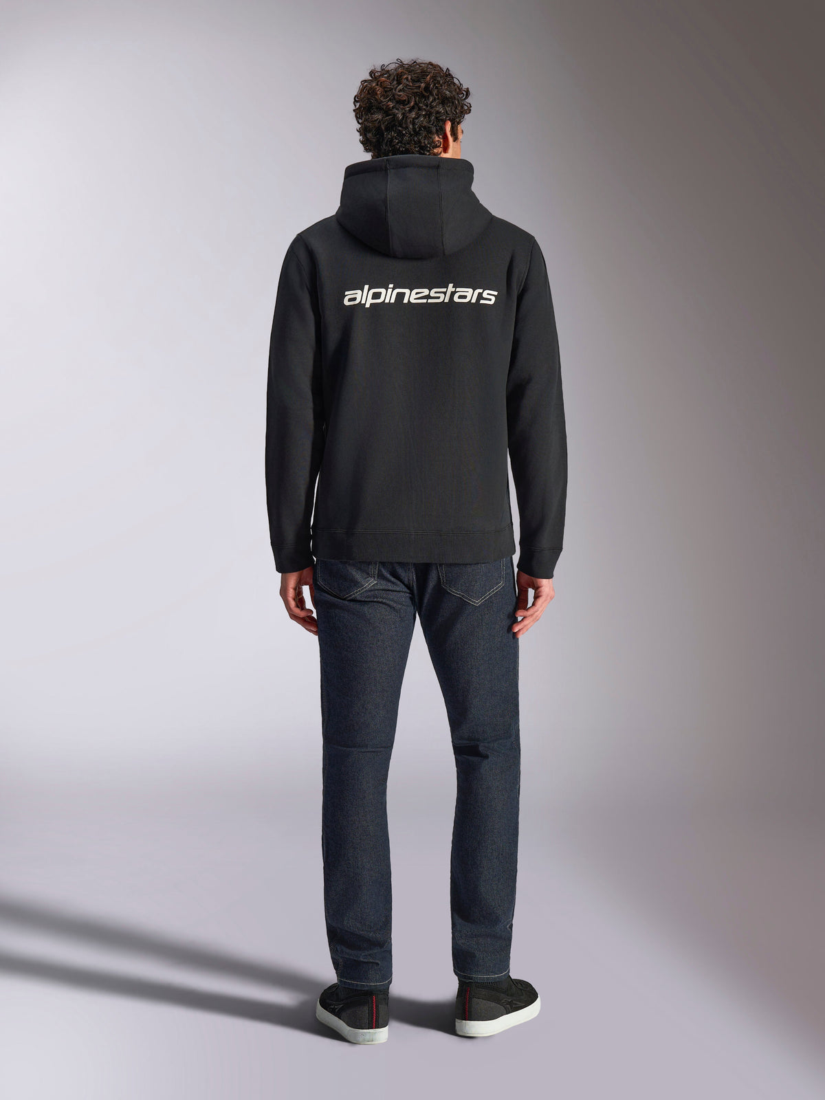 Alpinestars Transverse Hoodie, casual motorsport streetwear, zwart met wit Alpinestars-woordlogo op de rug, lange mouwen, geribde boorden en manchetten, van achteren gedragen met een donkere denim jeans