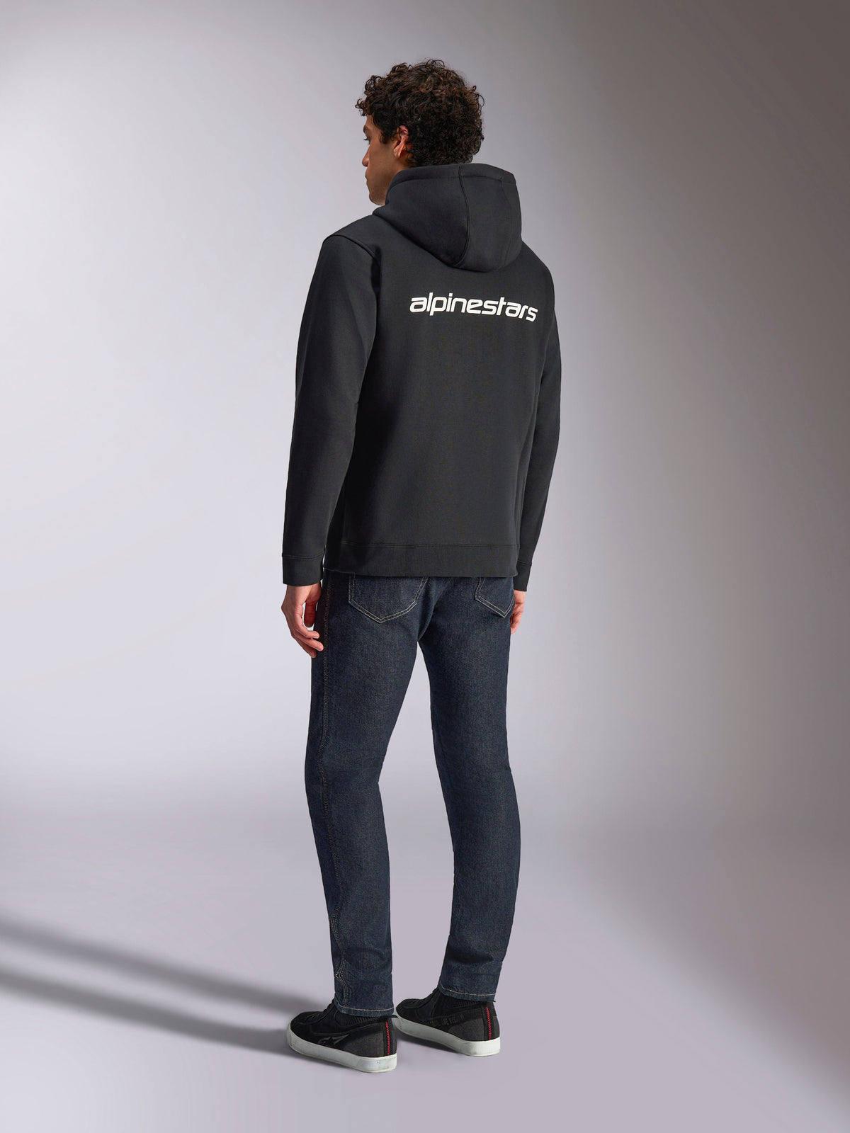 Alpinestars Transverse Hoodie, zwarte fleece hoodie met rits, achteraanzicht met een wit Alpinestars-woordlogo over de schouders, gedragen met donkerblauwe jeans en zwarte sneakers, casual op motorsport geïnspireerde streetwear