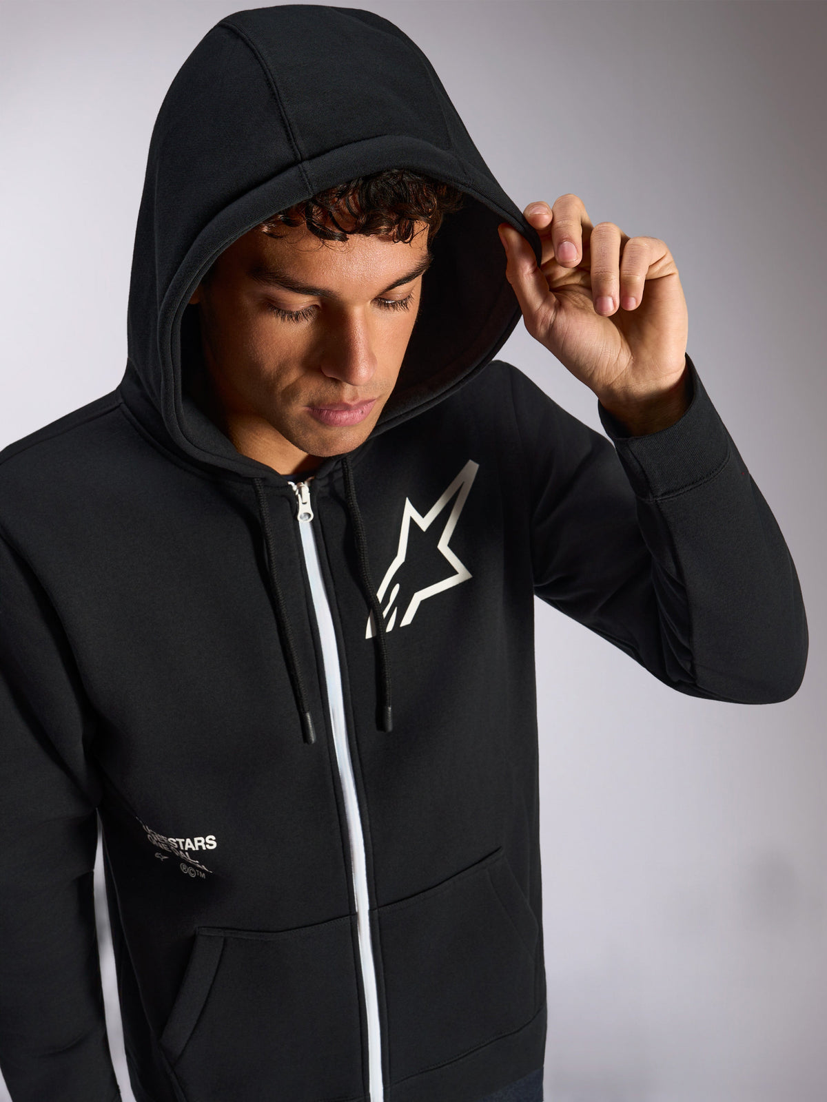 Alpinestars Transverse Hoodie, zwarte hoodie met ritssluiting aan de voorzijde, wit Astars-logo op de borst, witte rits, capuchon met trekkoord omhoog gedragen, casual motorsport-geïnspireerde streetwear