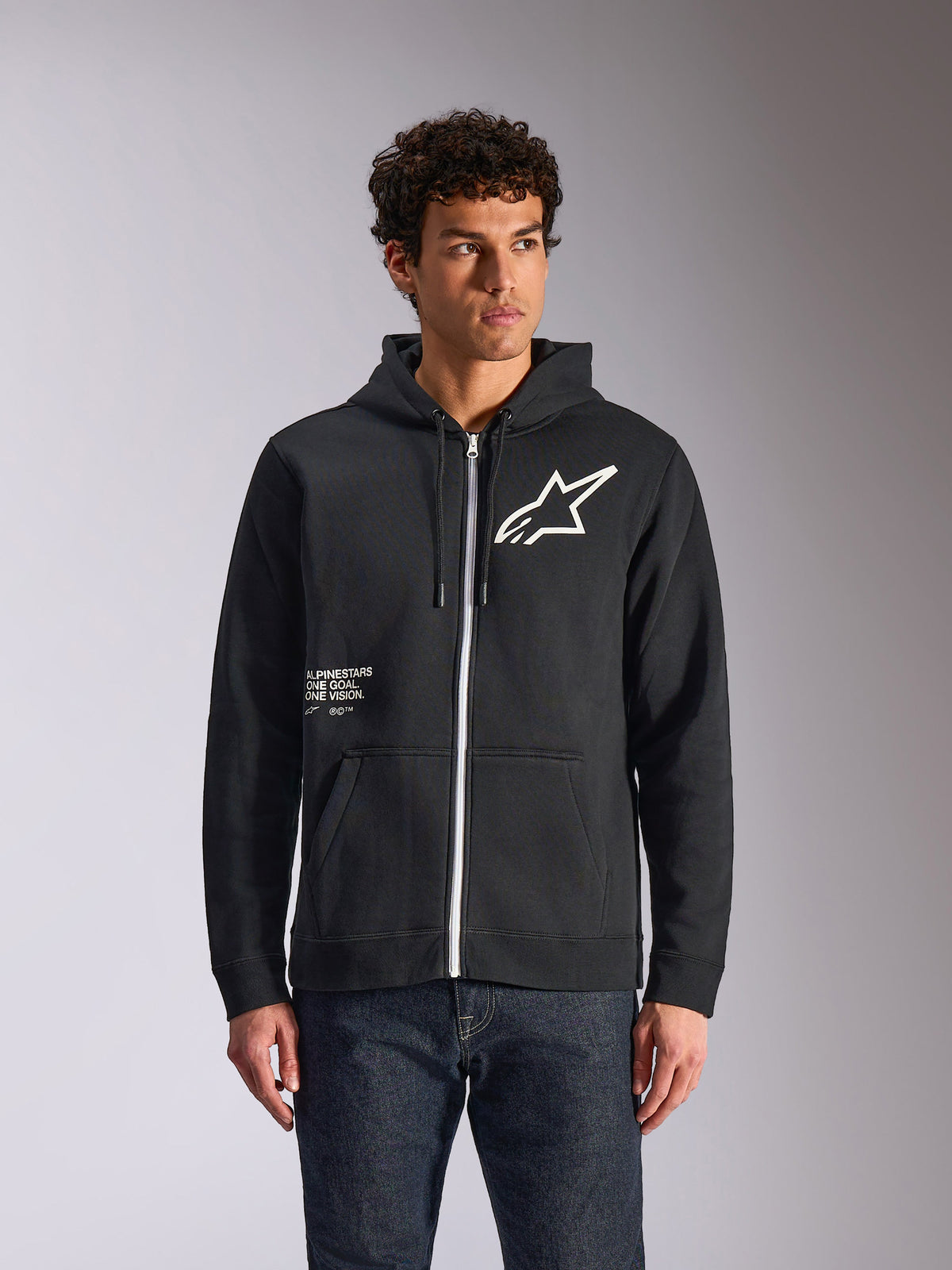 Alpinestars Transverse Hoodie, zwart fleecevest met rits en capuchon, voorzien van wit Astars-logo en One Goal-graphic op de voorzijde, gedragen door een model met een donkere jeans, casual motorsport-geïnspireerde streetwear