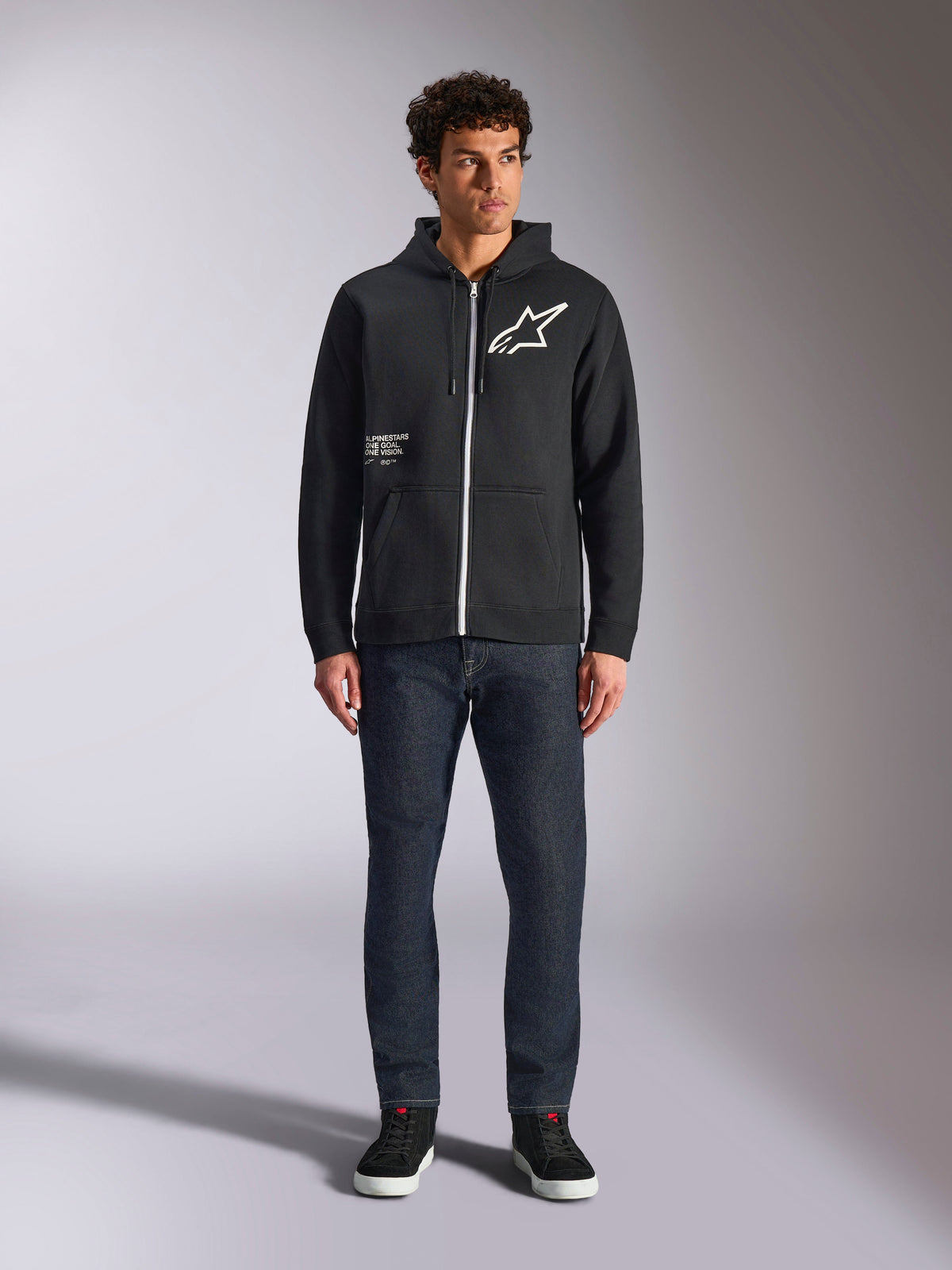 Alpinestars Transverse Hoodie, casual fleece hoodie met rits, zwart, wit Astars-logo op de borst en 'One Goal'-print aan de voorkant, capuchon met trekkoord, gecombineerd met een donkere jeans en zwarte sneakers, lifestyle motorsport streetwear