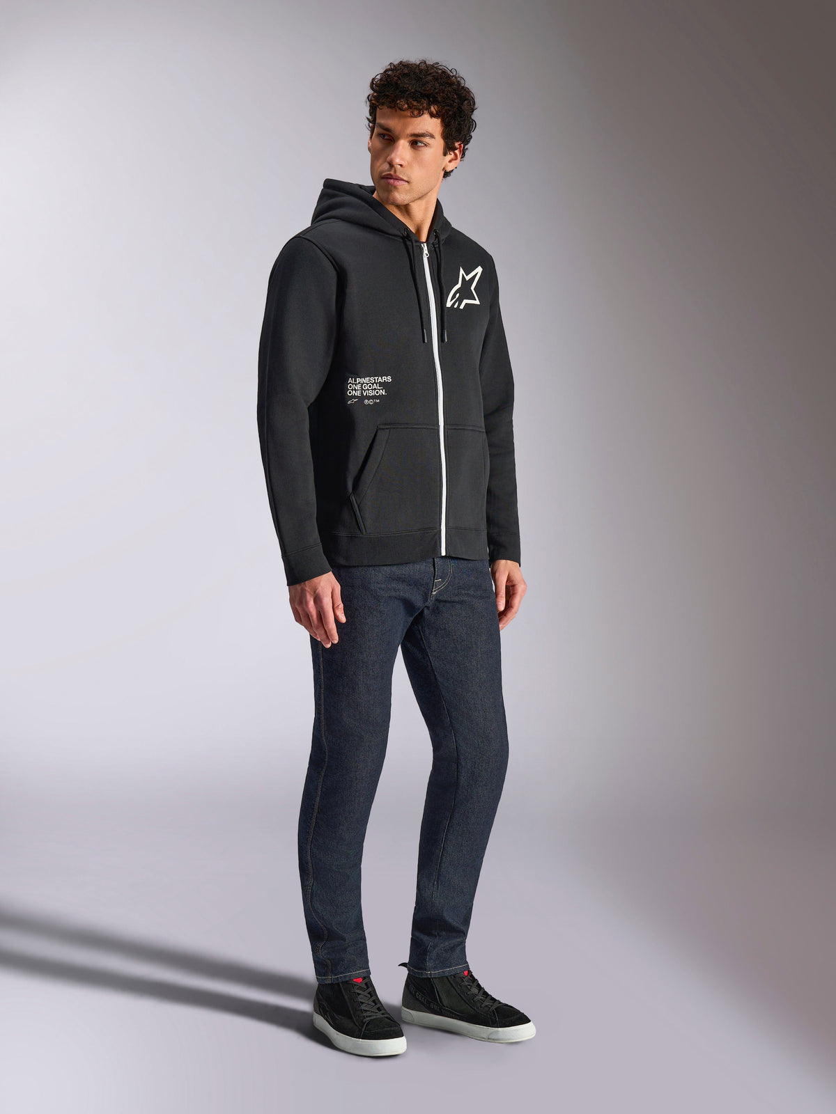 Alpinestars Transverse Hoodie, zwarte hoodie met rits, contrasterende witte rits, wit ster-logo op de borst en 'One Goal' branding op de zak, gedragen door een model met donkere jeans en zwarte sneakers, casual motorsport-geïnspireerde streetwear.