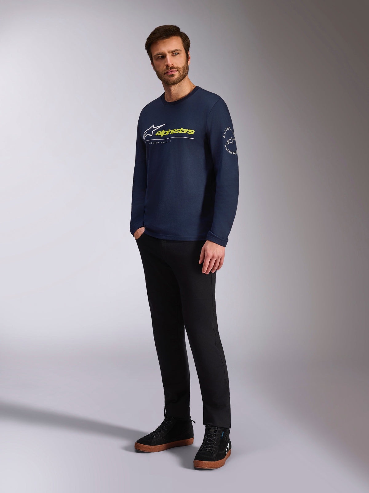 Alpinestars Mandatory CSF Tee, T-shirt met lange mouwen, navy, met wit Astars-logo en gele Alpinestars-branding op de voorzijde, Astars-cirkellogo op de mouw, gedragen door een model met zwarte broek en zwarte sneakers voor een casual lifestyle-look
