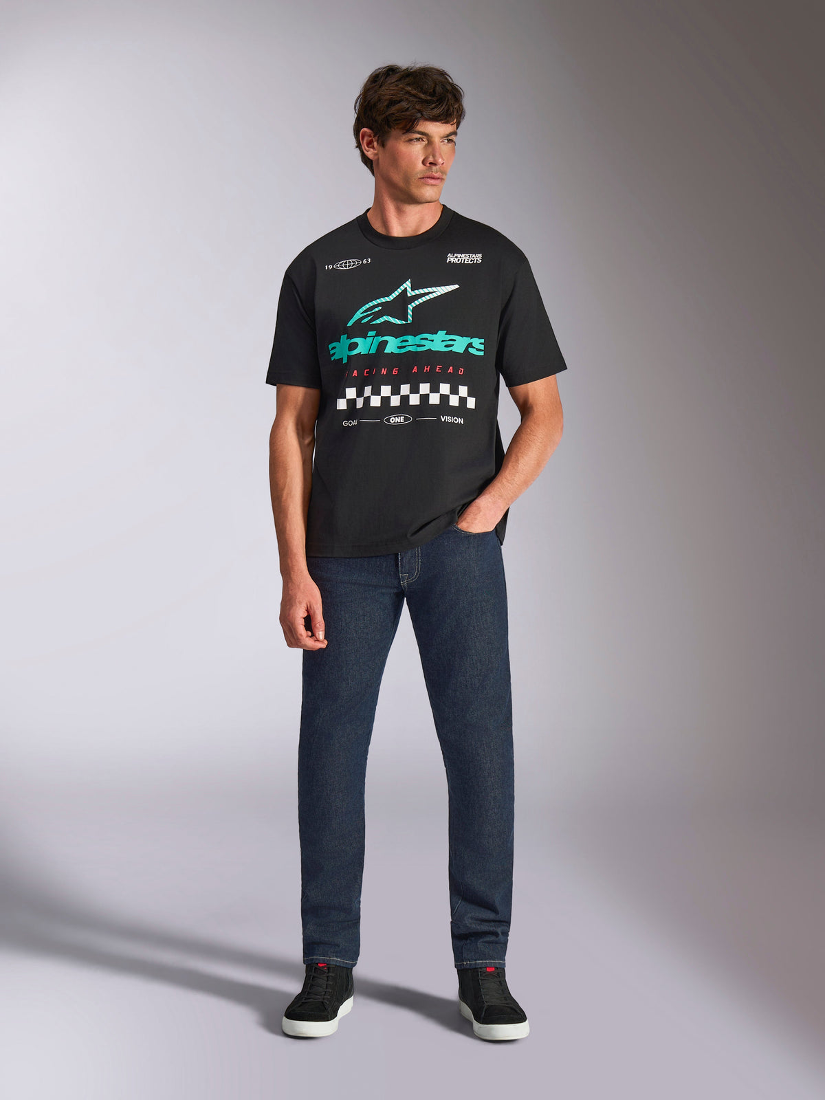 Alpinestars Aplomb Oversized CSF Tee, Short Sleeve T-Shirt, Black, Teal gradient Astars logo with red 'Racing Ahead' text and checkered details, worn by model with dark blue jeans, casual motorsport streetwear