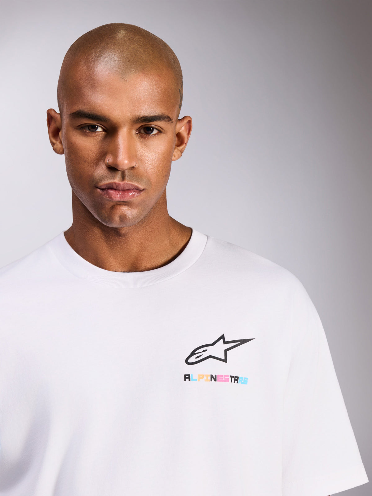 Mannelijk model draagt Alpinestars Wild Oversized CSF Tee, wit T-shirt met korte mouwen, voorzien van zwart Astars-logo met levendige meerkleurige letters op de borst, casual streetwear-stijl