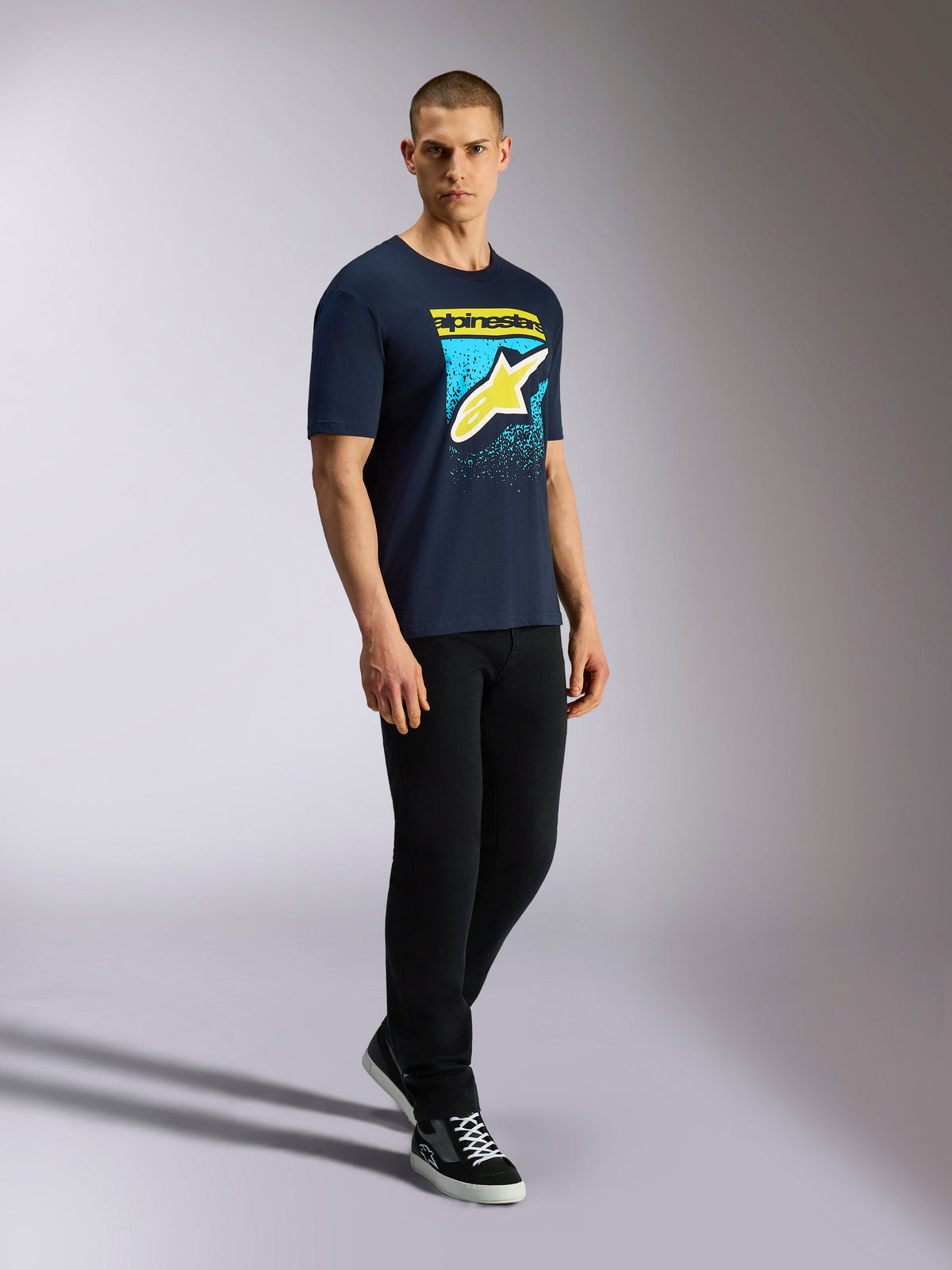 Alpinestars Noka CSF Tee, Short Sleeve T-Shirt, Navy, Yellow Astars Logo with Blue MX Gradation Graphic, Male model wearing navy tee with black pants and sneakers, Casual Motorsport Streetwear