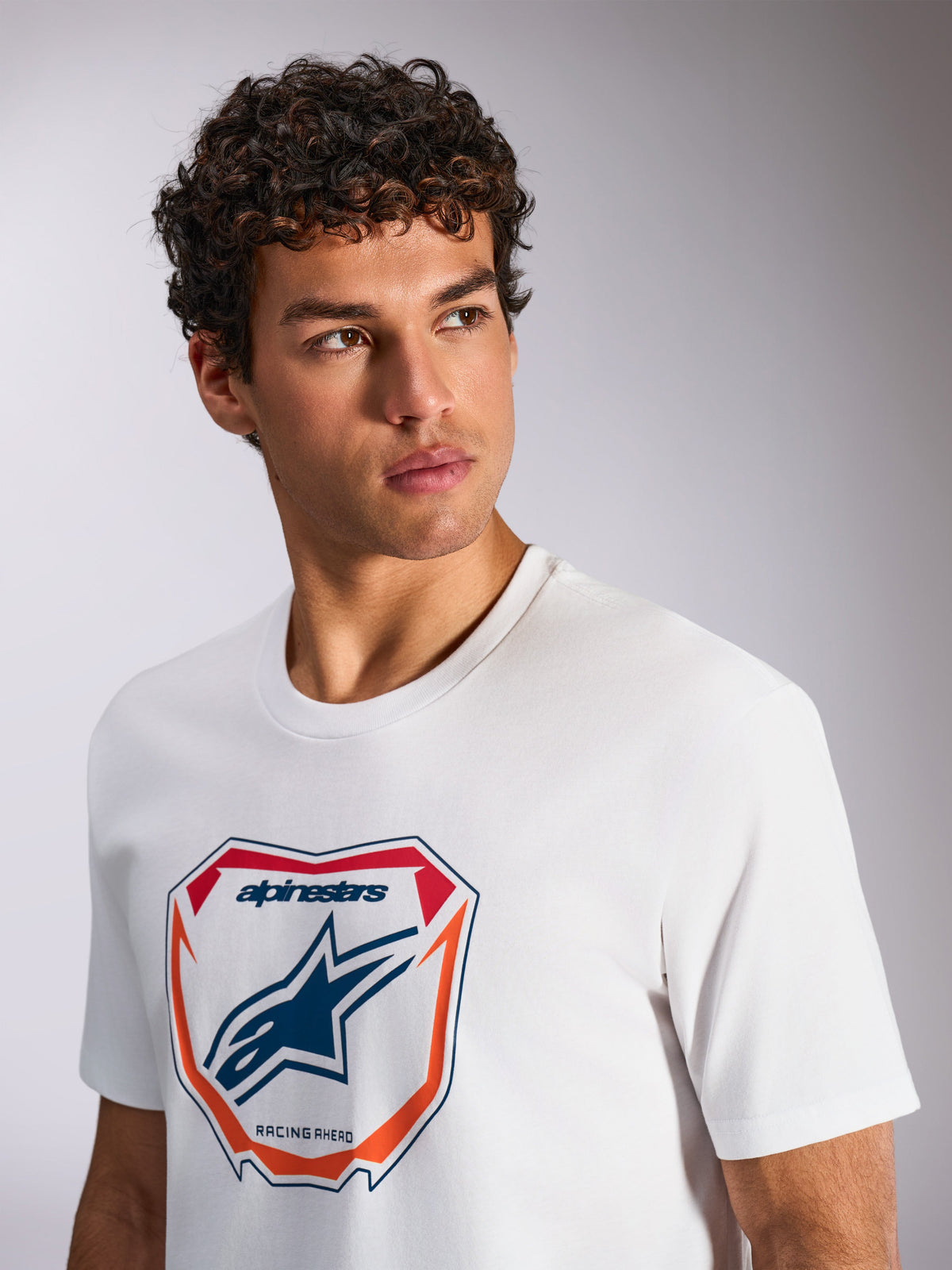 Alpinestars Plate CSF Tee, T-shirt met korte mouwen, wit, met een Astars MX plaatgrafiek op de voorzijde met rode en oranje accenten, 'Racing Ahead' tekst, gedragen door een model, casual motorsport streetwear