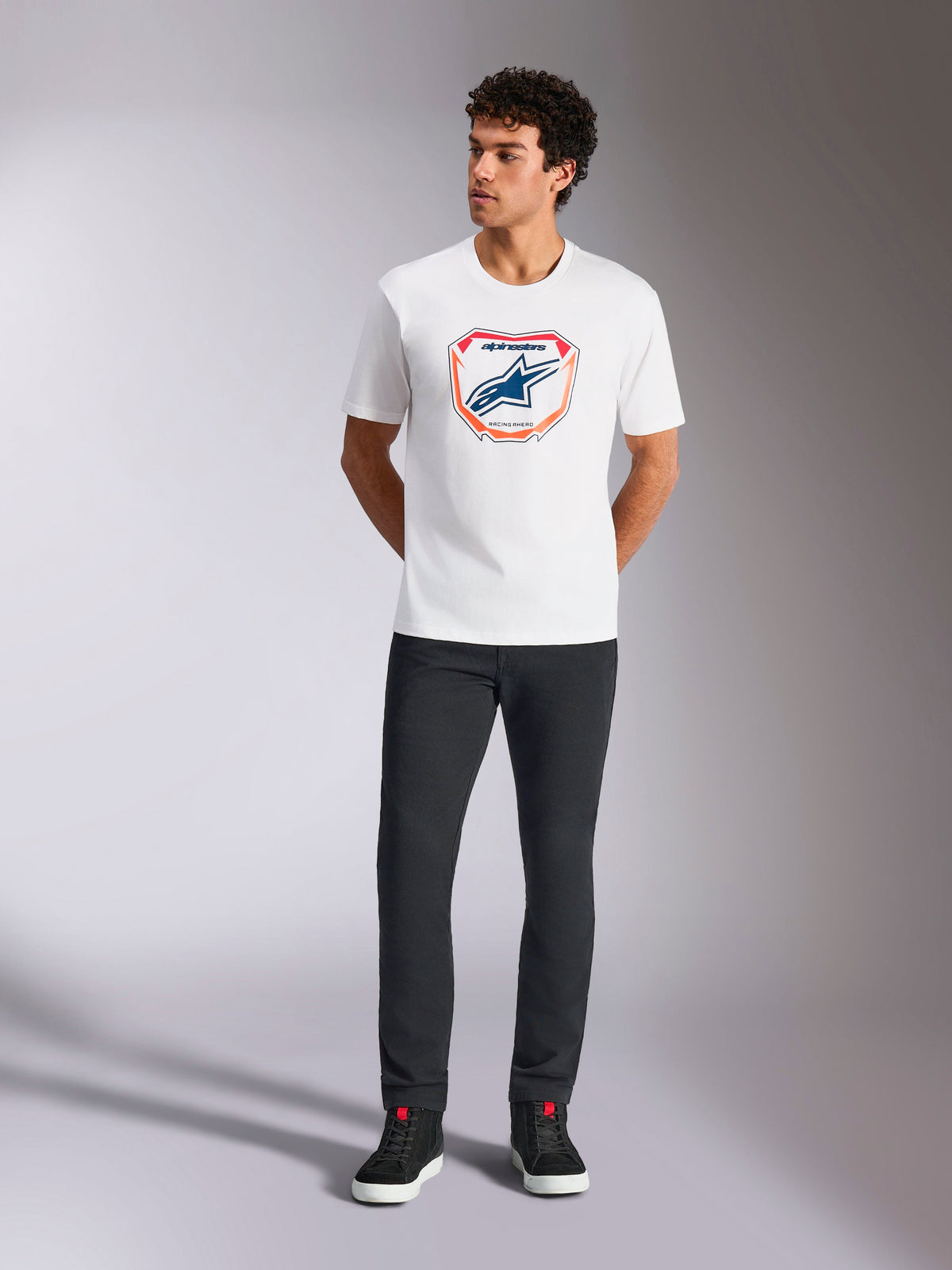 Alpinestars Plate CSF Tee, T-shirt met korte mouwen, wit, met een Astars MX plaatgrafiek aan de voorzijde met oranje en blauwe accenten, gedragen door een model met een zwarte broek en sneakers, casual motorsport streetwear