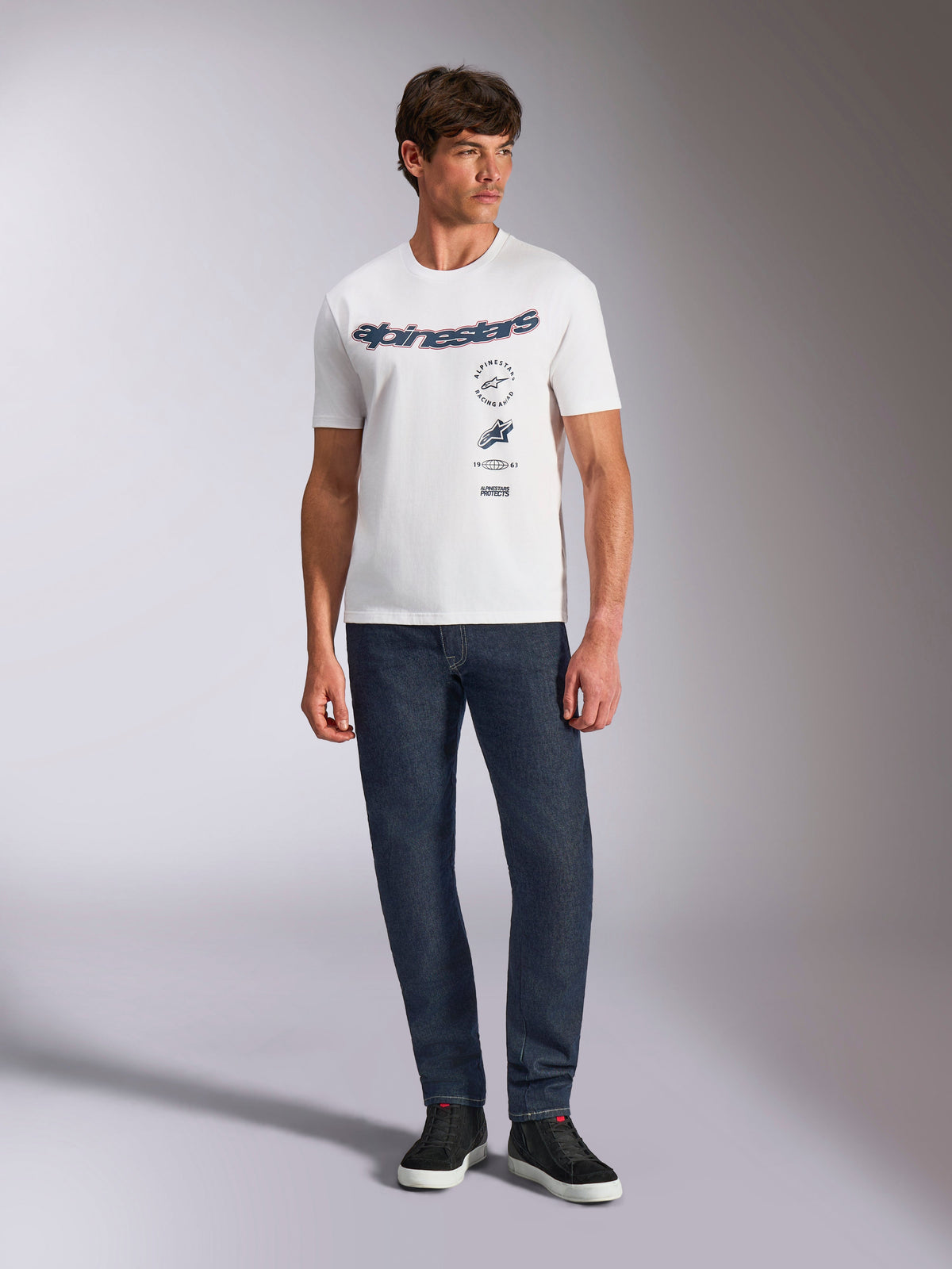 Alpinestars Abus CSF Tee, T-shirt met korte mouwen, wit, met gebogen Alpinestars-logo en meervoudige gestapelde graphics op de borst, gedragen door een mannelijk model met donkerblauwe jeans en zwarte sneakers, casual motorsport streetwear