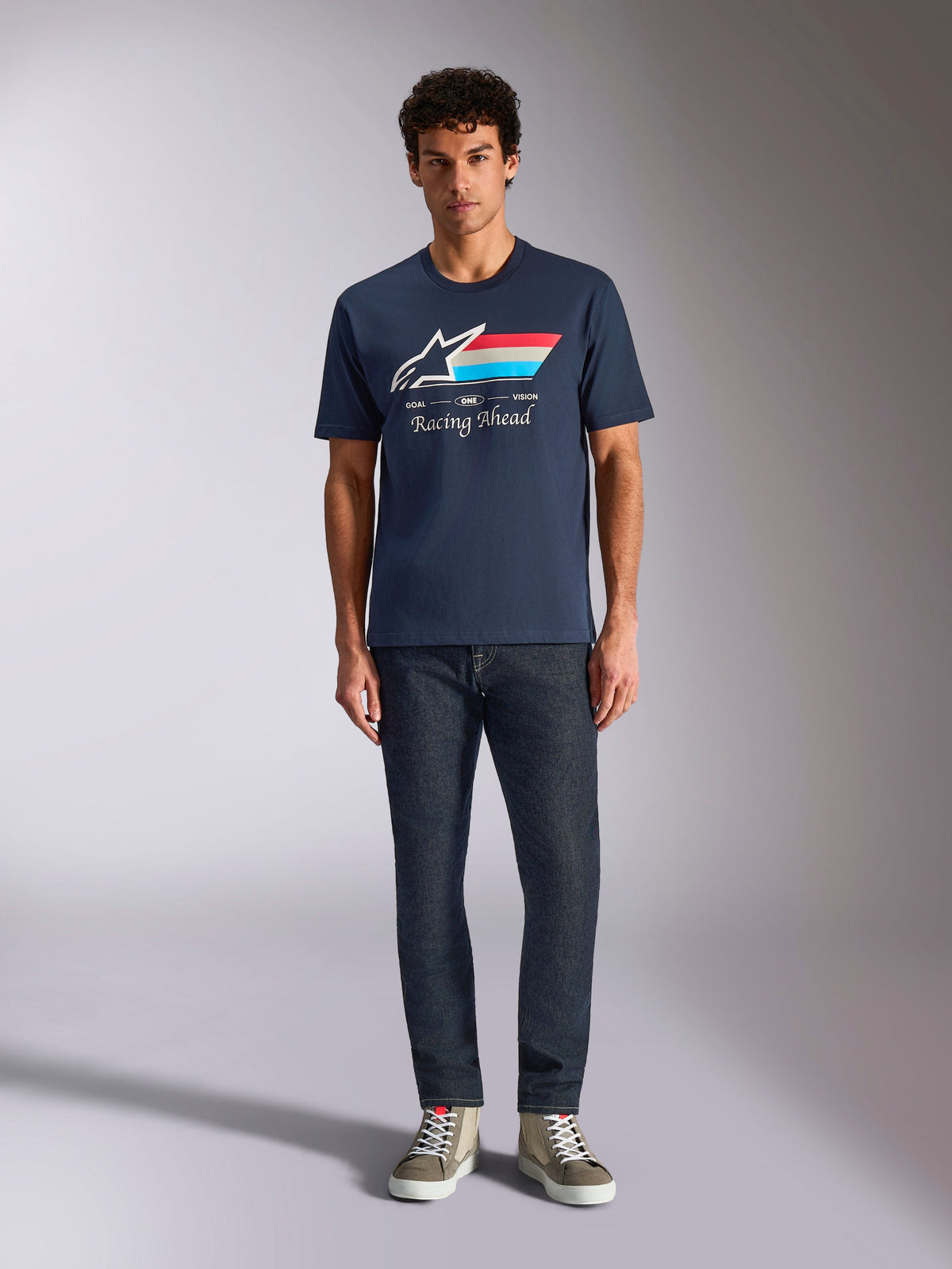 Alpinestars Trifecta CSF T-shirt met korte mouwen, marineblauw, wit Astars-logo met rode, witte en lichtblauwe strepen en 'Racing Ahead' tekst, gedragen met donkere jeans en olijfgroene sneakers, casual motorsport streetwear