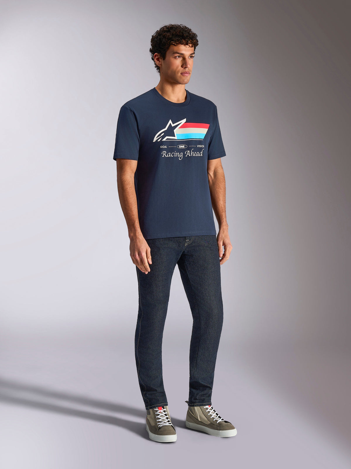 Alpinestars Trifecta CSF Tee, T-shirt met korte mouwen, marineblauw, voorzien van Astars-logo met driekleurige strepen en 'Racing Ahead'-tekst, gedragen door een model met donkere jeans en sneakers, casual door motorsport geïnspireerde streetwear