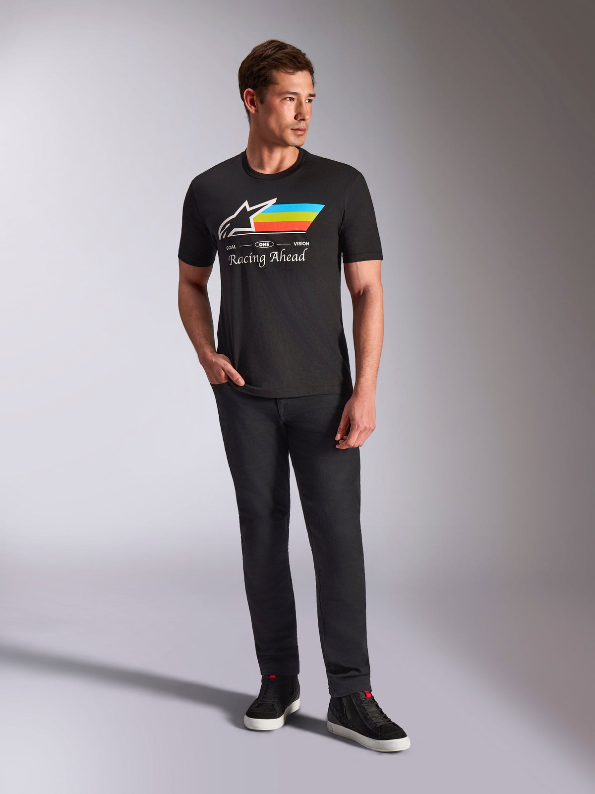 Alpinestars Trifecta CSF Tee, Short Sleeve T-Shirt, Black, featuring a white Astars logo with cyan, yellow, and orange stripes and 'Racing Ahead' text, worn by a model with black pants and sneakers for casual motorsport streetwear