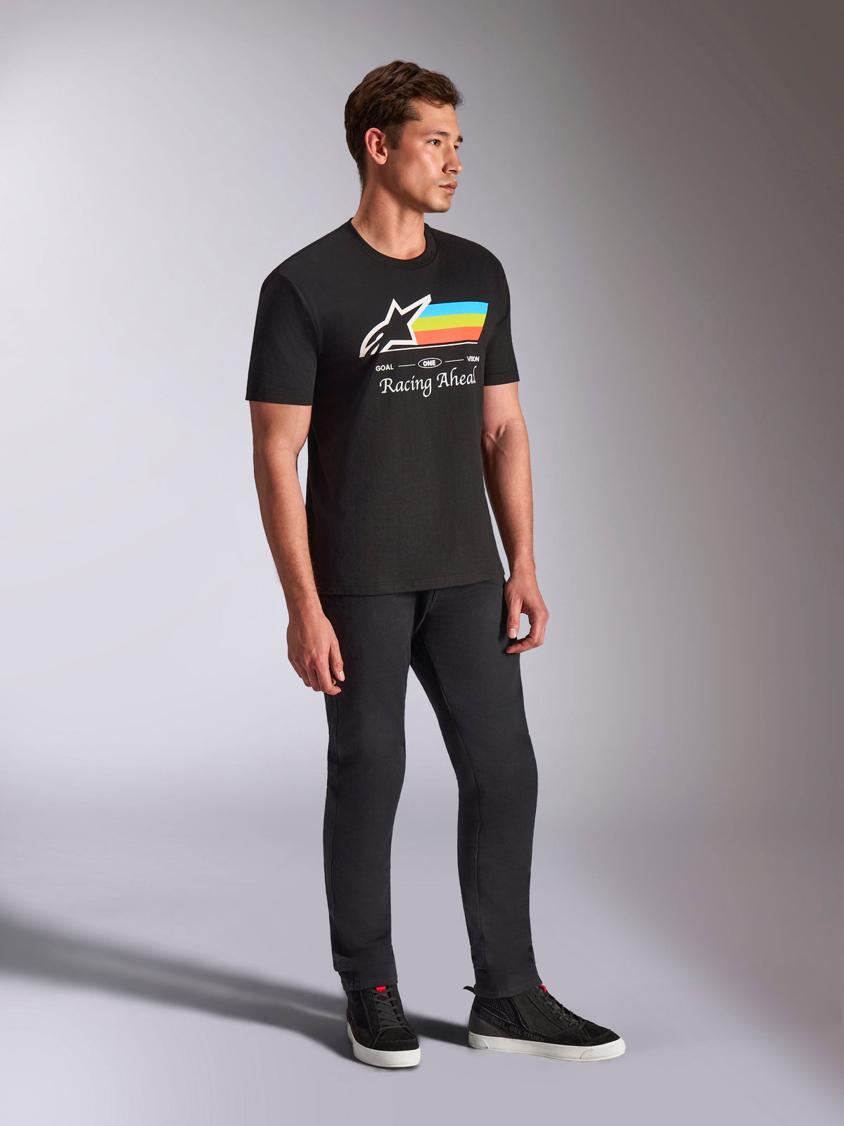 Alpinestars Trifecta CSF Tee, Short Sleeve T-Shirt, Black, white Astars logo with blue, yellow, and orange stripes and 'Racing Ahead' graphic, worn by a model with black pants and sneakers, casual motorsport-inspired lifestyle apparel